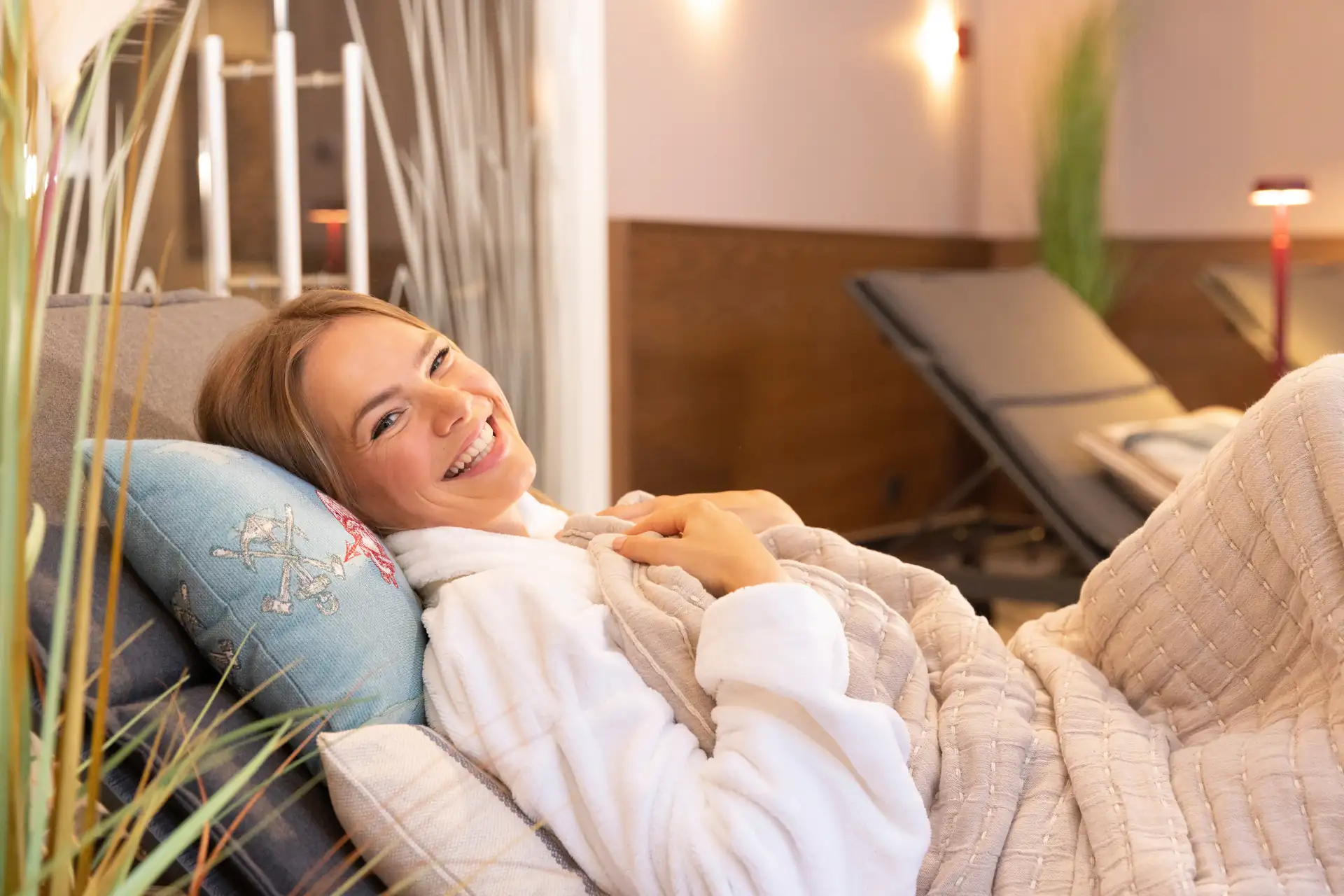 Hotel City Krone: Eine Frau liegt entspannt auf einer Liege, eingekuschelt in eine Decke. Sie trägt einen weißen Bademantel und hat eine Kissen mit einem blauen Bezug. Im Hintergrund sind Liegen und Pflanzen sichtbar.
