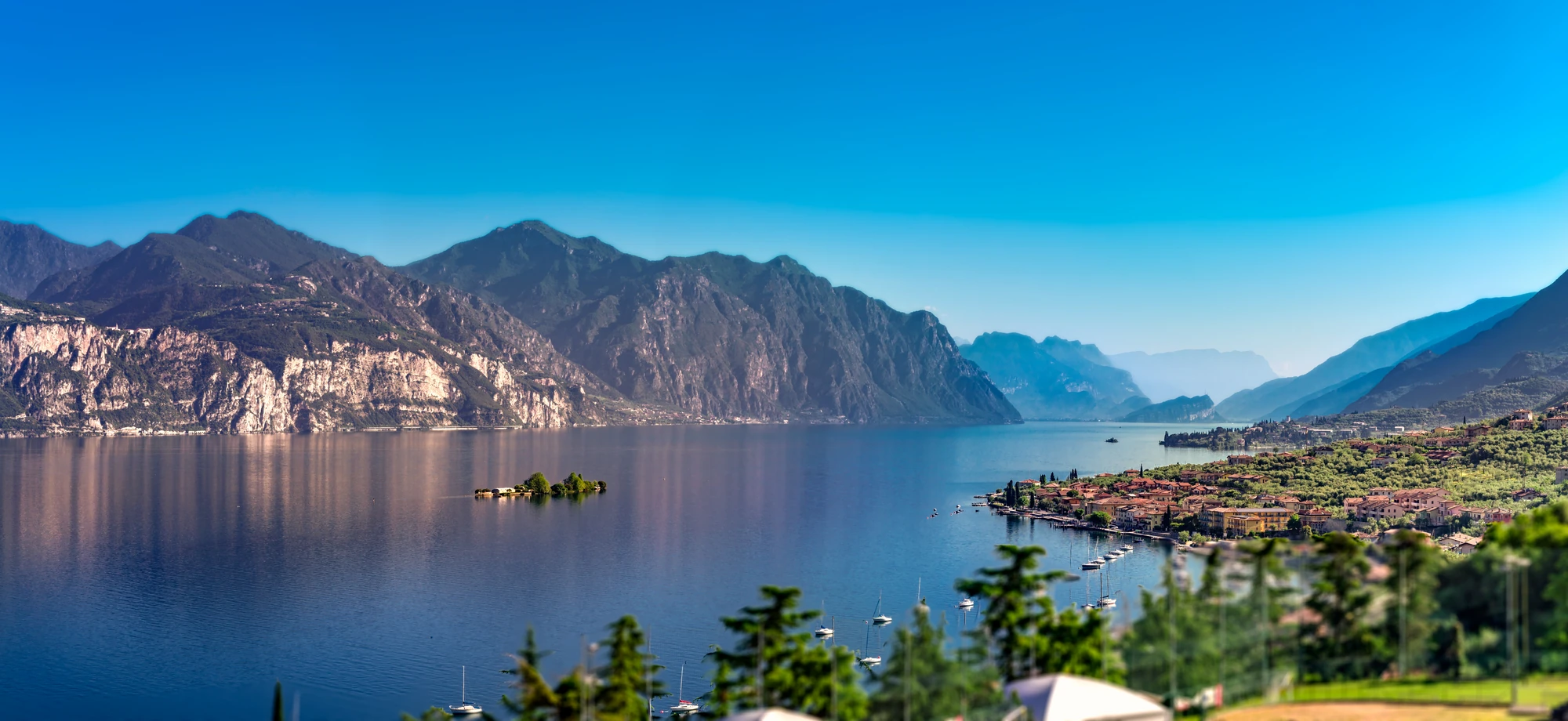Blick auf den glitzernden Gardasee, mit den majestätischen Bergen links und bunten Häusern rechts, die von üppiger Natur umgeben sind. Bei sonnigem Wetter strahlt die Szene eine lebendige und einladende Atmosphäre aus.