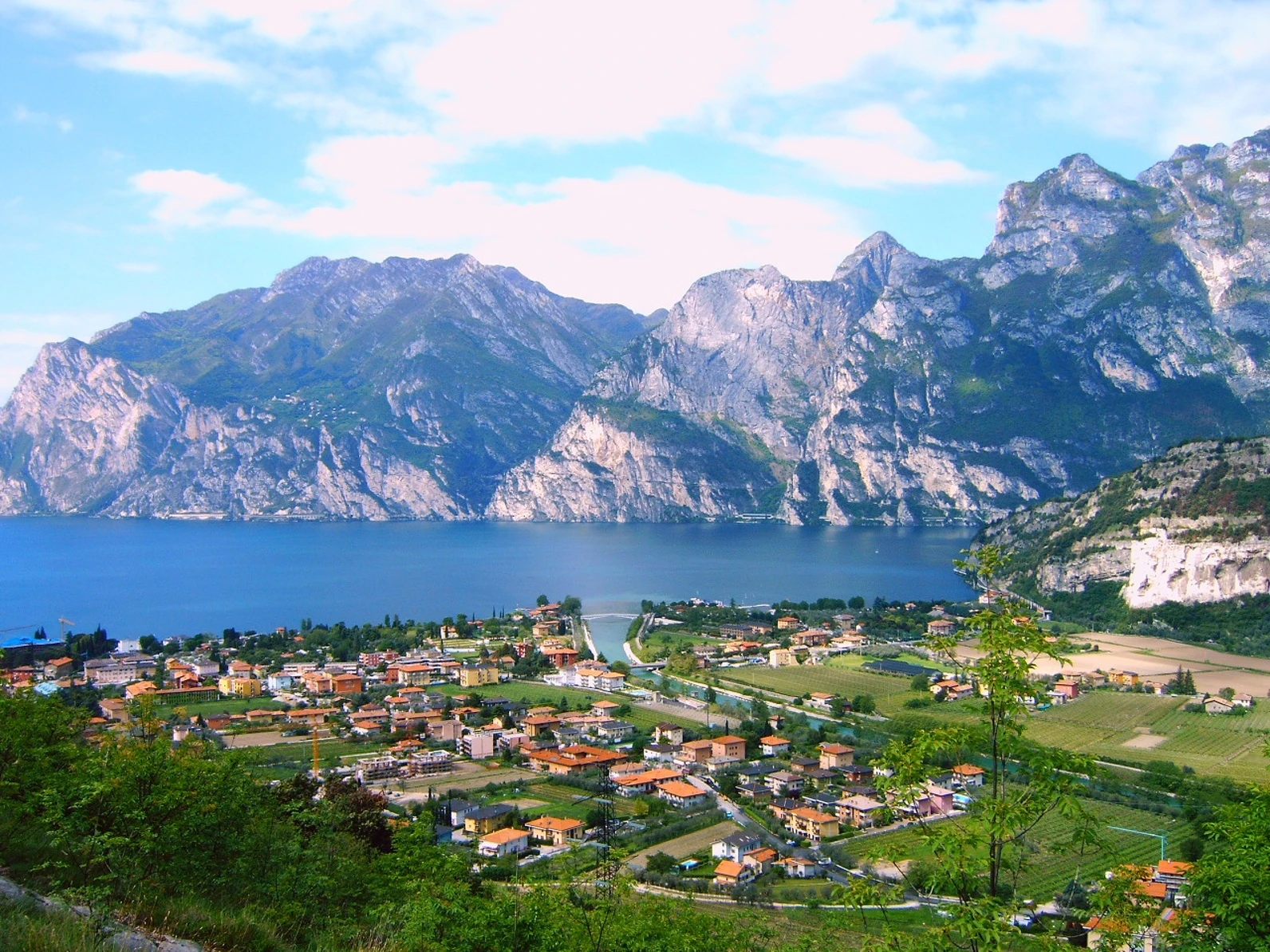Panoramablick über den Gardasee, der die charmanten Häuser zeigt, umgeben von üppiger Natur. Das glitzernde Wasser des Sees spiegelt die umliegenden Berge wider, was eine beeindruckende und harmonische Landschaft schafft.