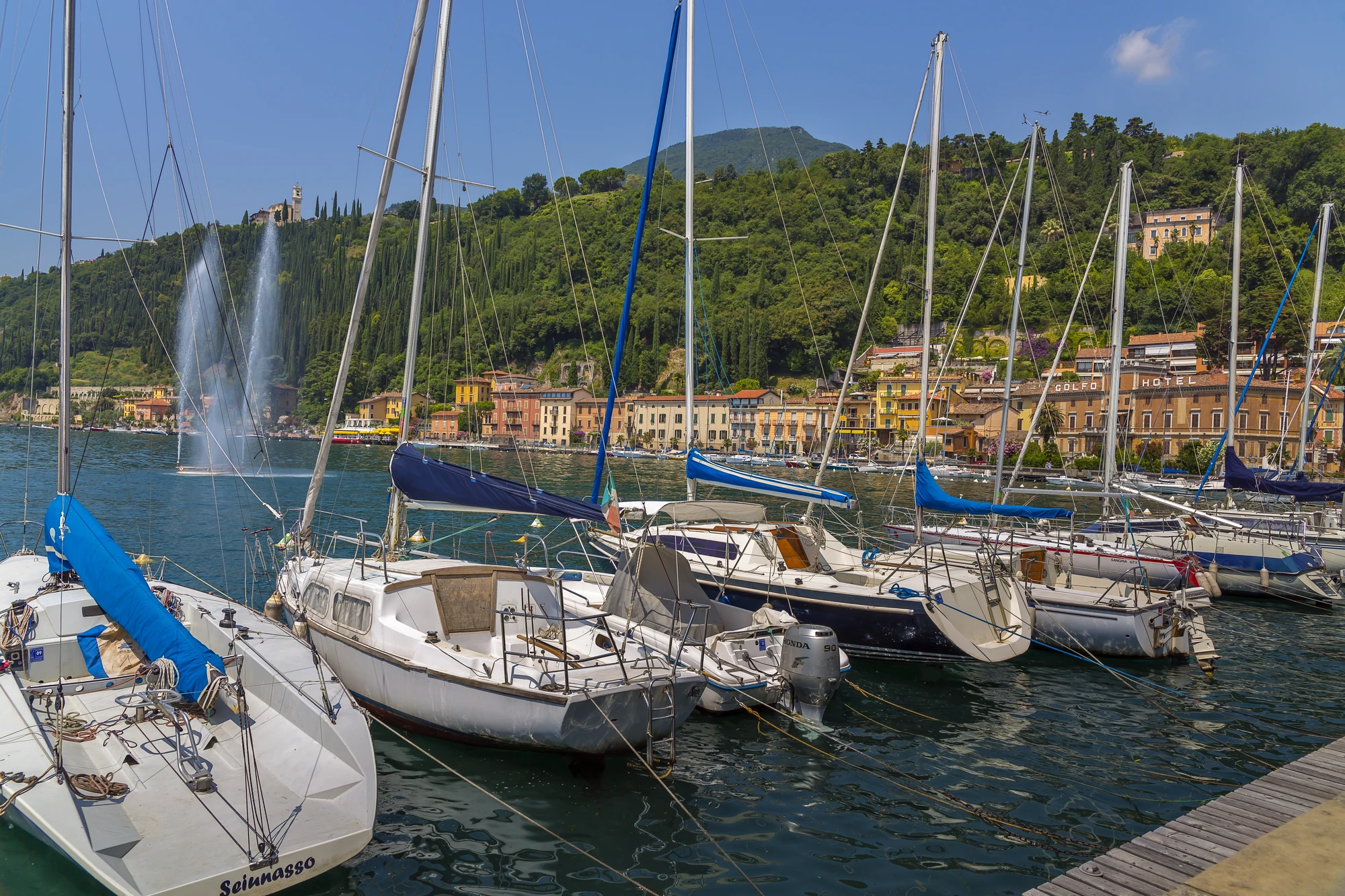 Boote im Hafen am Gardasee, umgeben von bunten Häusern an der Promenade. Im Hintergrund erstrecken sich majestätische Berge und üppige Bäume unter klarem Wetter. Die lebendige Kulisse und das glitzernde Wasser schaffen eine einladende und malerische Atmosphäre.