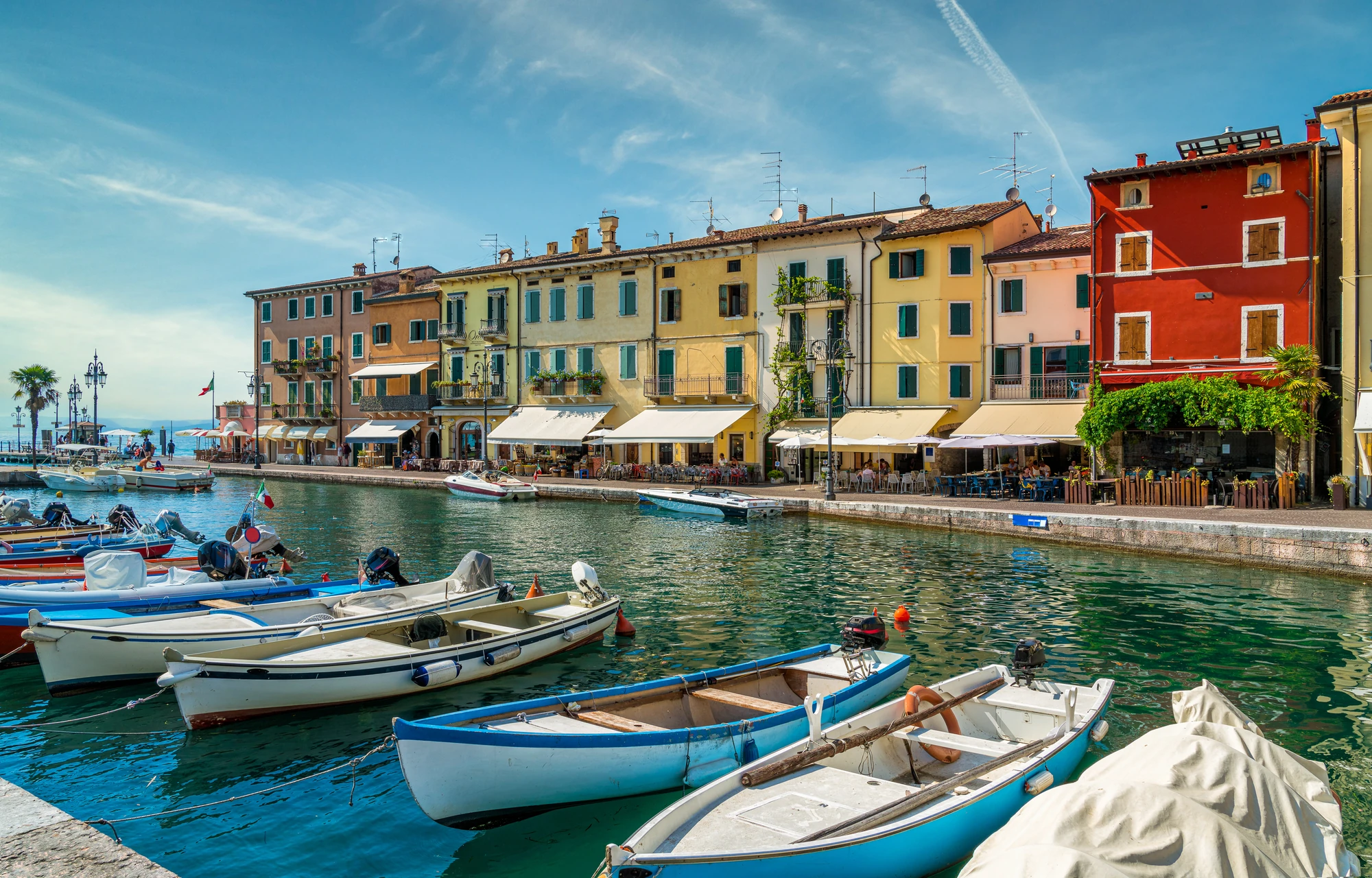 Hafen in Lazise am Gardasee, umgeben von bunten Häusern und kleinen Booten, die sanft im Wasser schaukeln. Bei schönem sonnigen Wetter strahlt die Szene eine lebendige und einladende Atmosphäre aus, während die Farben der Gebäude das malerische Ambiente unterstreichen.