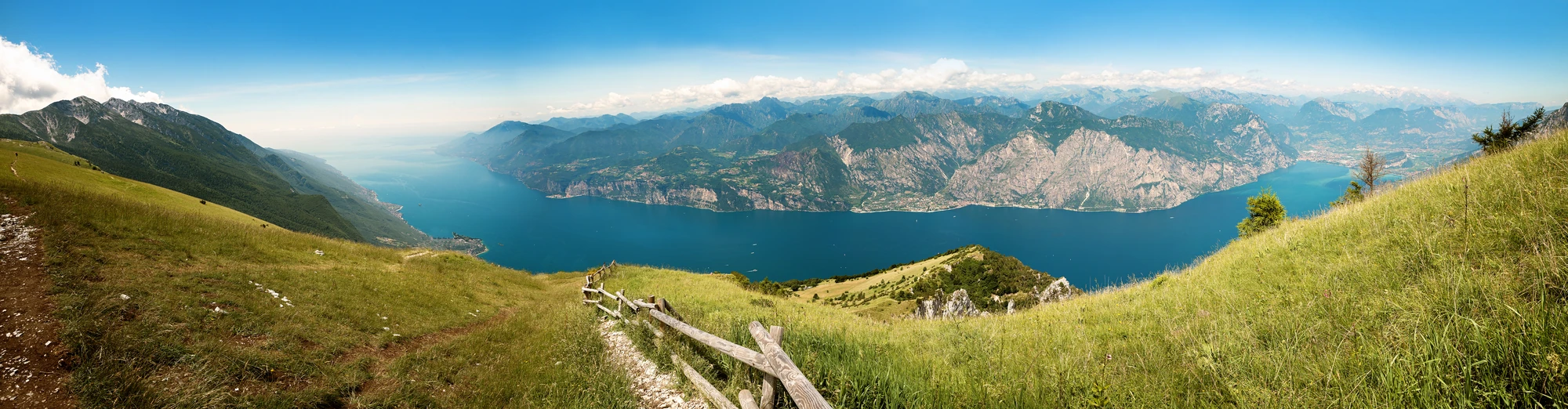 Panoramablick vom Monte Baldo auf den Gardasee und die umliegenden Berge. Die beeindruckende Landschaft bietet eine atemberaubende Aussicht auf das glitzernde Wasser des Sees, umrahmt von grünen Hügeln und schroffen Gipfeln, die in der Ferne emporragen.