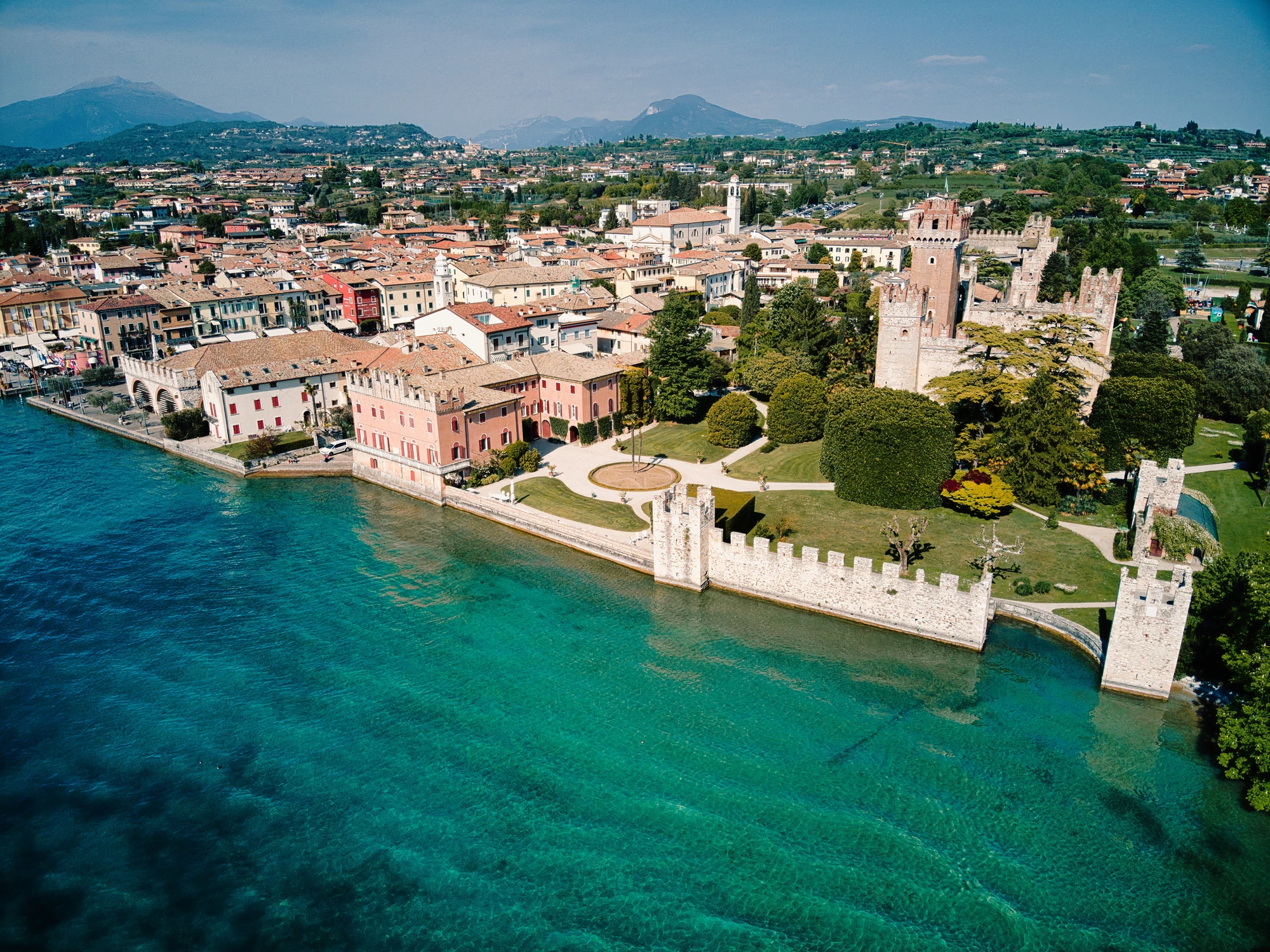 Panoramafoto über die Stadt Lazise am Gardasee, mit Fokus auf die beeindruckende Scaliger-Burg und die Villa des Grafen Bernini. Die malerische Küstenlinie und das glitzernde Wasser des Sees ergänzen die historische Architektur und schaffen eine harmonische Atmosphäre.
