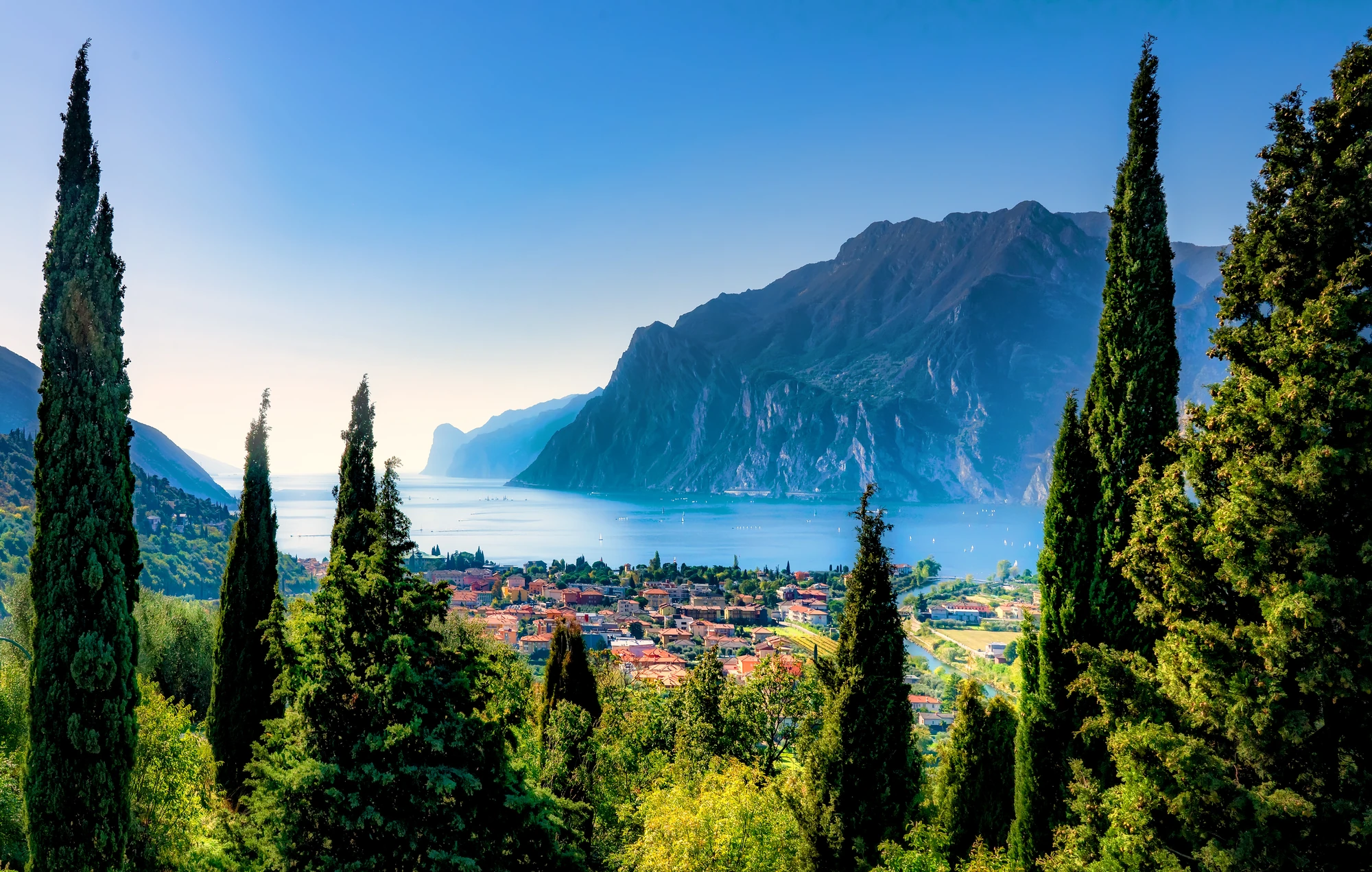 Blick auf Torbole am Gardasee durch die Bäumen hindurch, mit der Stadt im Vordergrund und den beeindruckenden Bergen im Hintergrund. Diese Perspektive schafft eine malerische und natürliche Rahmung der idyllischen Landschaft.