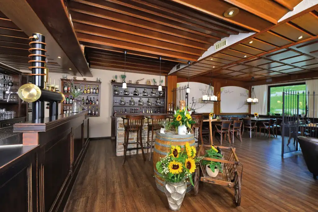 Holzgetäfelte Weinstube mit Bar, Holzbalkendecke, Holzboden, Tischen und Stühlen sowie einem mit Sonnenblumen dekorierten Weinfass in der Mitte