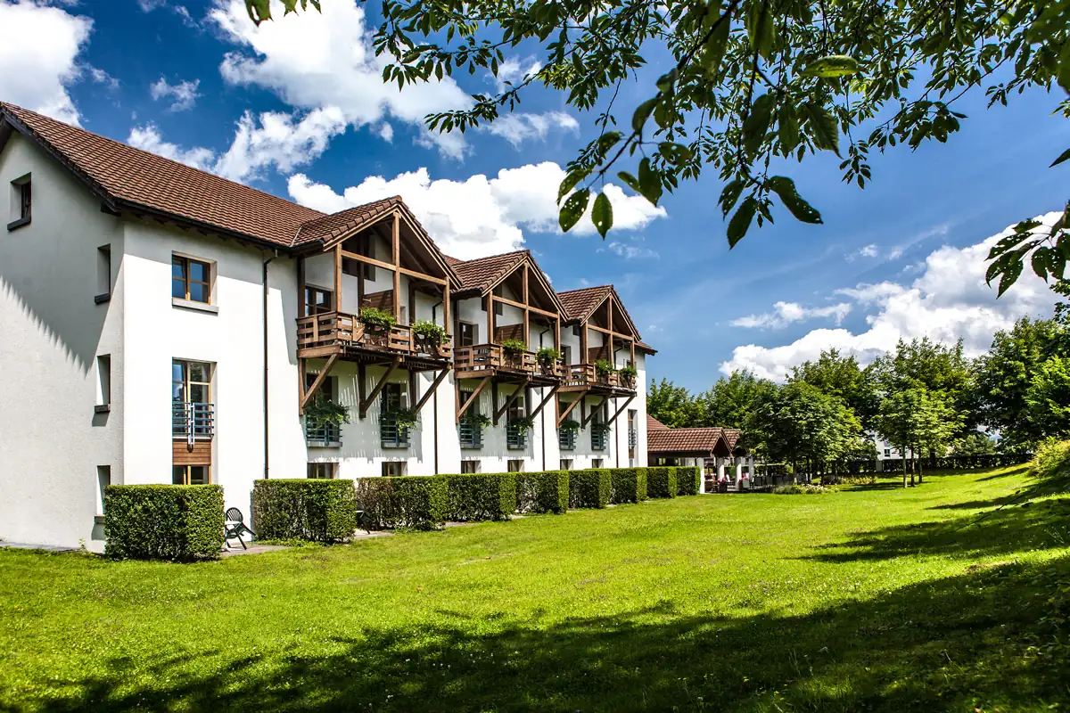 Hotel Seegarten in Arbon: Mehrstöckiges weißes Hotelgebäude mit Holzbalkonen und roten Ziegeldächern, umgeben von grünem Rasen und Bäumen unter blauem Himmel mit Wolken, Beispielhotel für die Radreise Bodensee