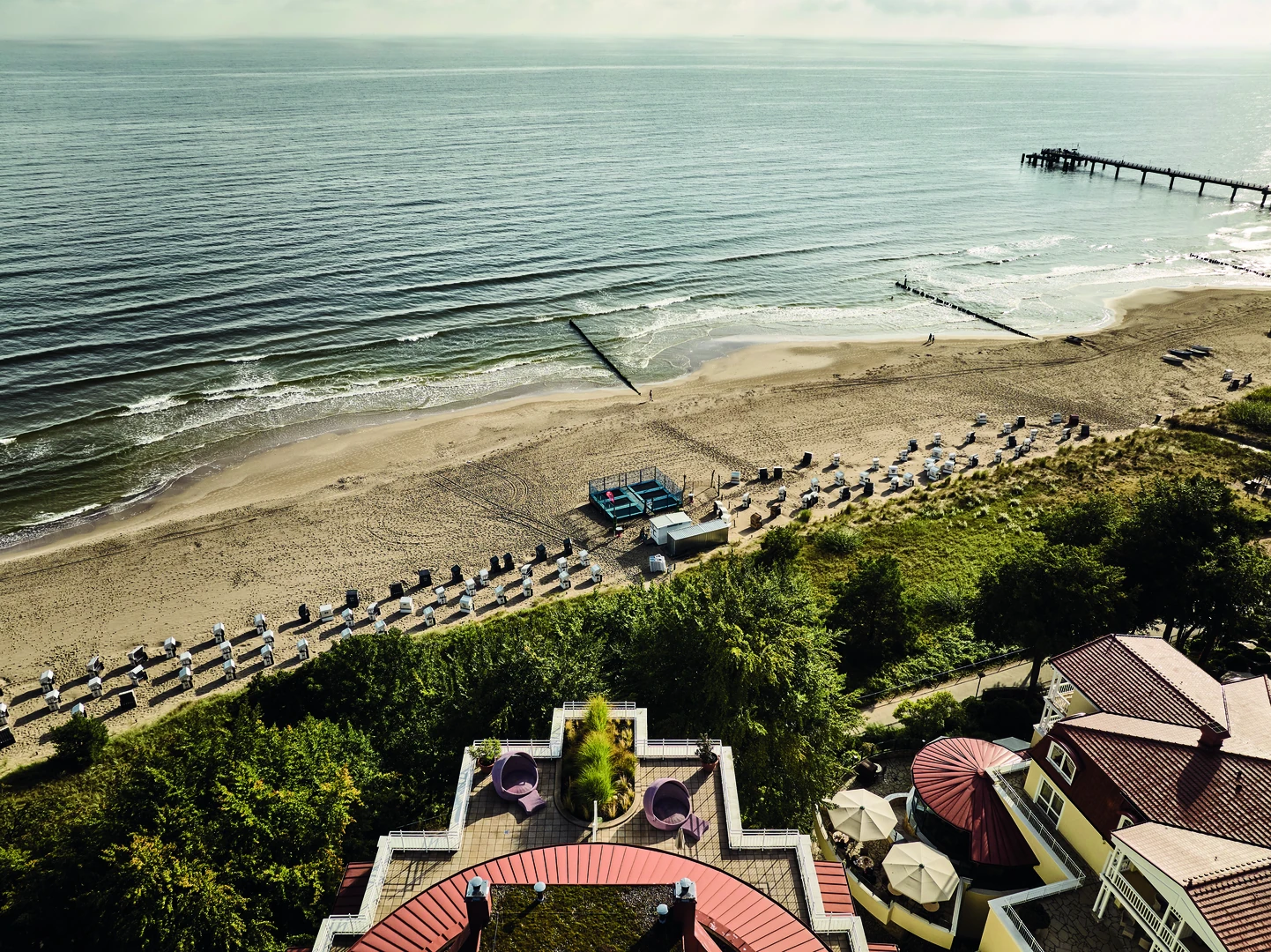 Ein beeindruckender Ausblick vom aja Strandhotel Bansin: Die Vogelperspektive zeigt den feinsandigen Strand der Ostsee, gesäumt von Strandkörben und sanften Wellen. Die idyllische Lage direkt am Meer verspricht Erholung und Entspannung, während die gepflegte Hotelanlage mit ihren Terrassen einladend wirkt. Ein perfekter Ort für einen unvergesslichen Urlaub.