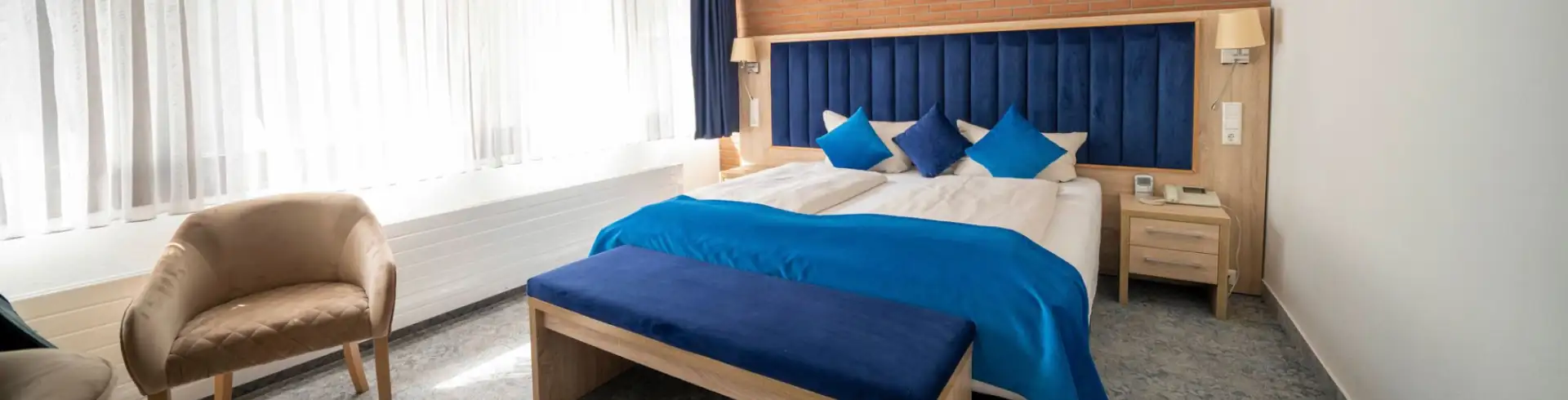 Doppelzimmer im Hotel Zur Heidschnucke mit großem Bett, blauer Bettdecke und Kissen, Holzbank am Fußende, zwei Sesseln und großen Fenstern mit weißen Vorhängen (im Bannerformat)
