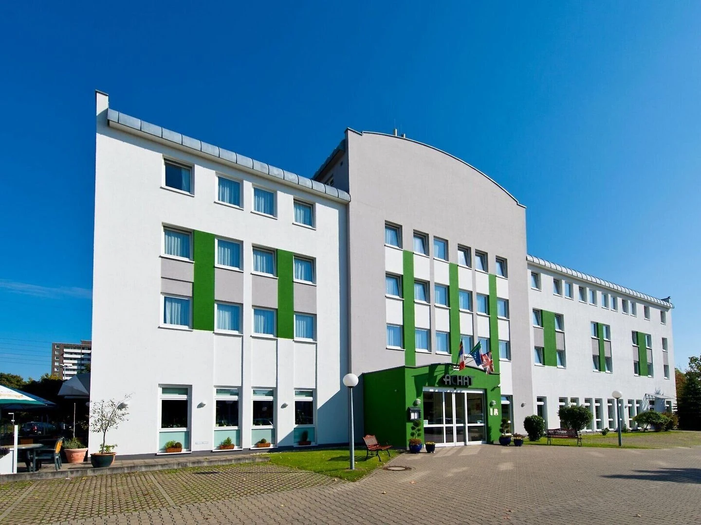 Dreigeschossiges Hotelgebäude mit weißer Fassade und grünen Fensterläden, Eingangsbereich mit grünem Vordach und Schriftzug 'ACHAT'
