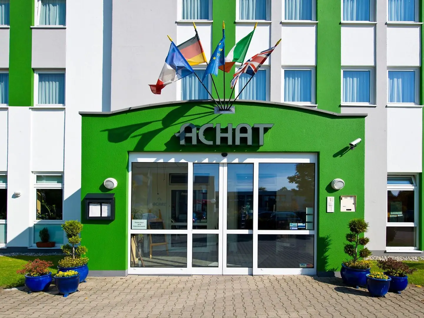 Eingang des ACHAT Hotels Monheim am Rhein mit grüner Fassade, Glastür, mehreren Flaggen und blauen Pflanzgefäßen mit kleinen Bäumen und Blumen