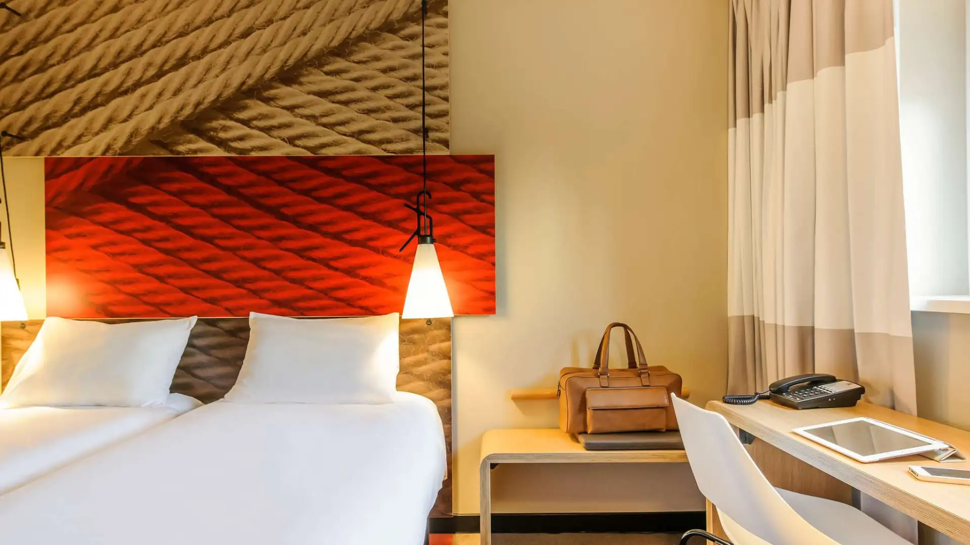 ibis Hotels im Kachelformat: Modernes Hotelzimmer im ibis Hamburg City mit Doppelbett, rotem Kopfteil, Schreibtisch mit Tablet und brauner Tasche.