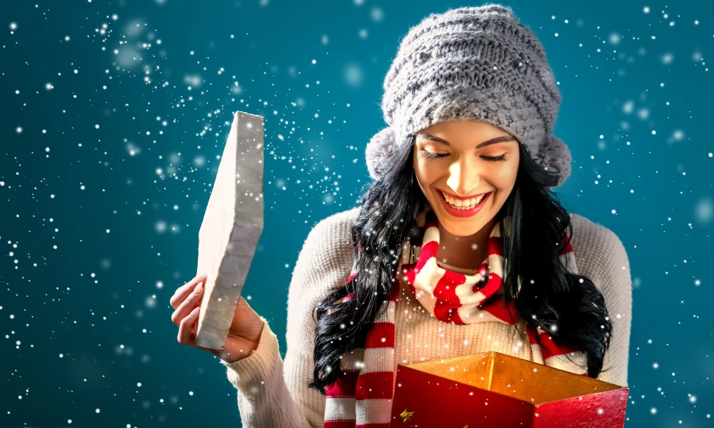 Das Bild zeigt eine junge Frau, die mit einem strahlenden Lächeln ein Weihnachtsgeschenk aus einer roten Schachtel betrachtet. Sie trägt eine graue Mütze und einen gestreiften Schal in Rot und Weiß. Der Hintergrund ist in weihnachtlichen Blautönen gehalten, während Schneeflocken um sie herum fallen, was eine festliche und gemütliche Atmosphäre schafft. Ihre Freude über das Geschenk wird deutlich durch ihr Lächeln und die begeisterte Mimik.