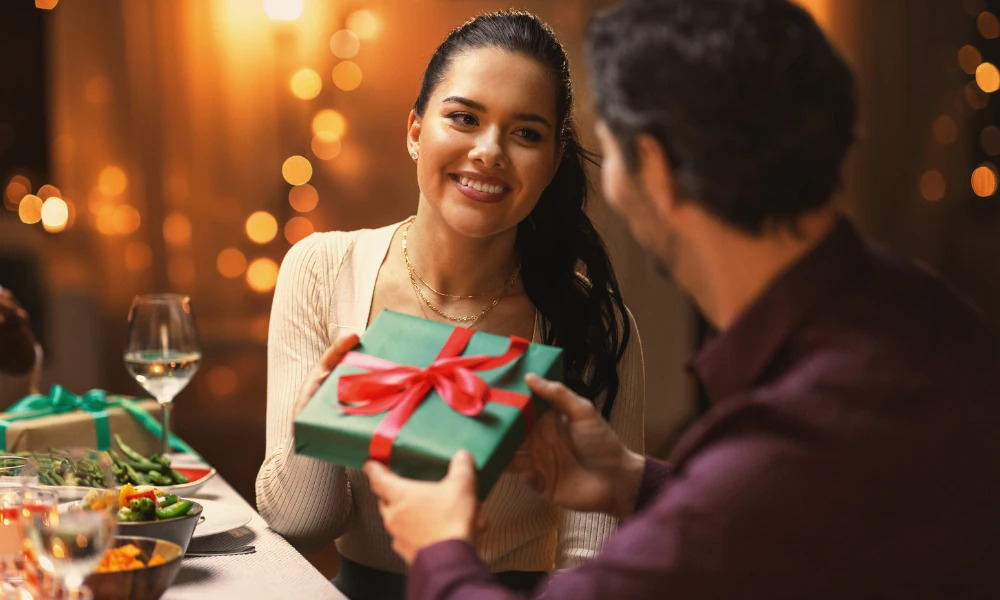 Das Bild zeigt ein Paar bei einer festlichen Mahlzeit, in einem warmen und einladenden Ambiente. Die Frau lächelt glücklich, während sie ein Geschenk in grüner Verpackung mit rotem Band hält. Sie übergibt es ihrem Partner, der sie ebenfalls lächelnd ansieht. Der Hintergrund ist mit weichen Lichtern dekoriert, die eine festliche Atmosphäre verstärken. Der Tisch ist gedeckt und zeigt Essen und Getränke, was auf eine gemütliche, intime Feier hindeutet.