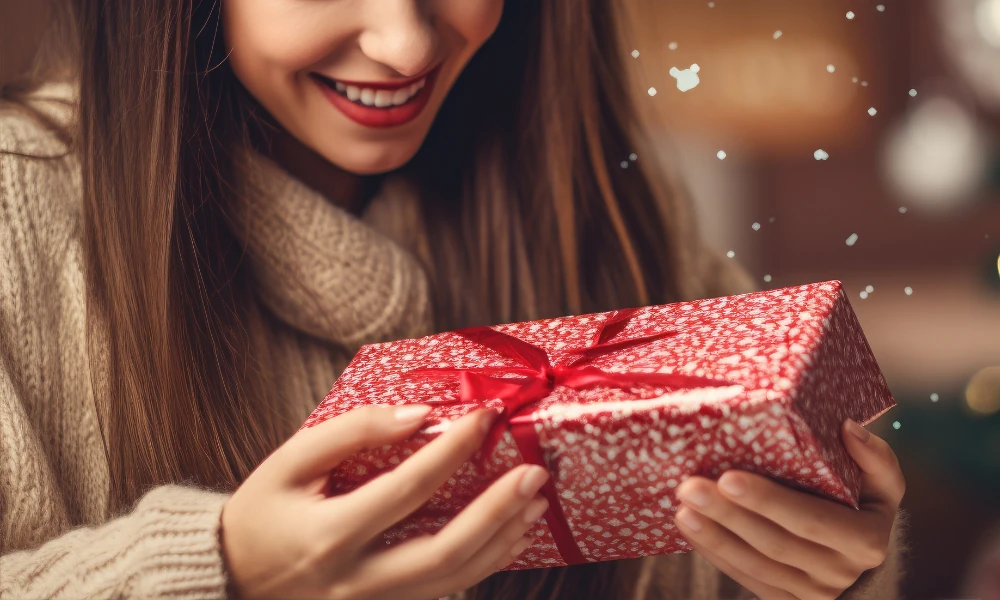 Das Bild zeigt eine Frau in einem warmen, beigen Pullover, die ein festlich verpacktes Geschenk mit rotem Band in den Händen hält. Sie lächelt, während sie das Geschenk betrachtet. Der Hintergrund ist unscharf und schafft eine gemütliche, weihnachtliche Atmosphäre, unterstützt von einem Hauch von Schneeflocken, die das Bild verschönern. Die Szene vermittelt Freude und Vorfreude auf die festliche Jahreszeit.