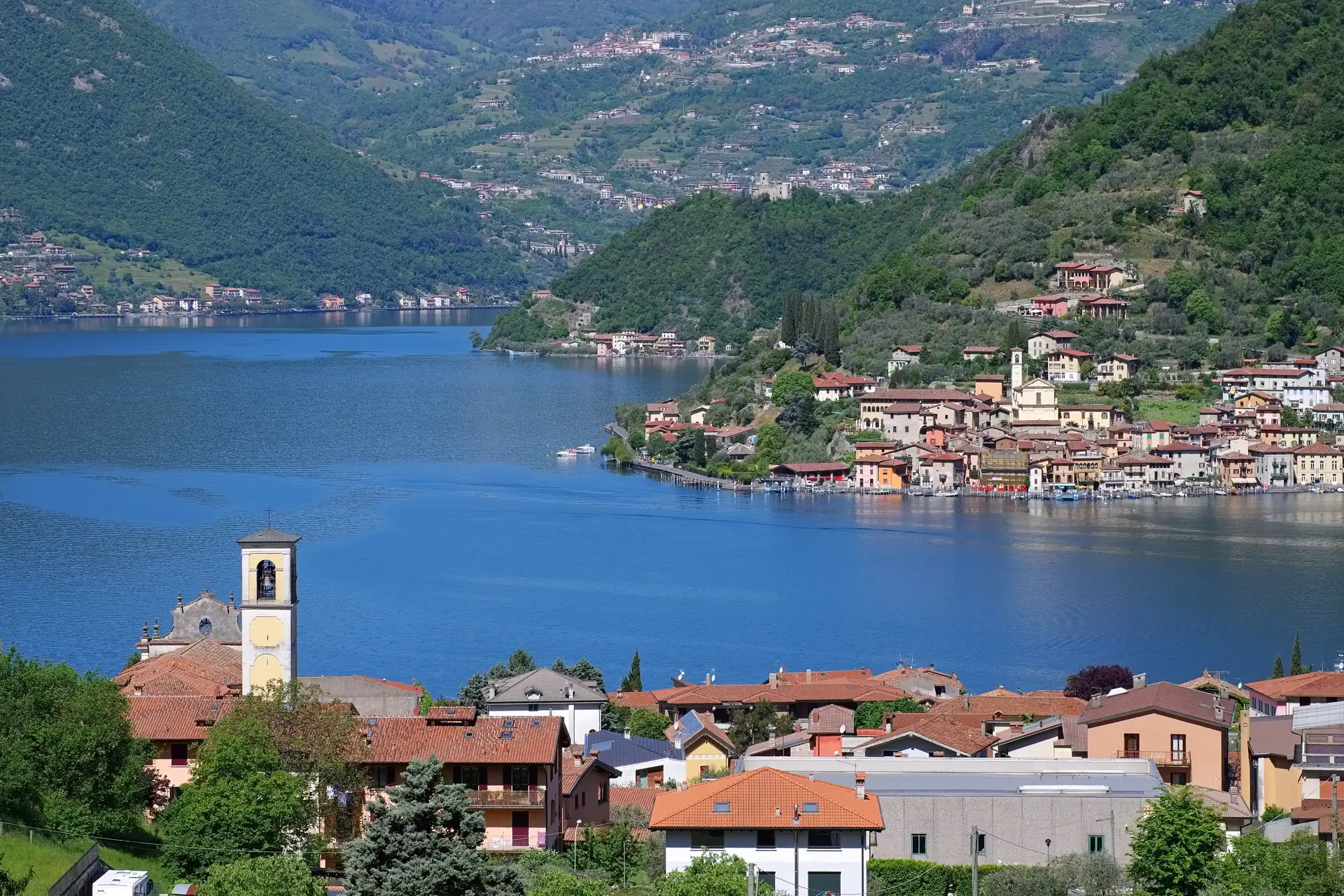 Blick auf den Iseosee mit der Insel Monte Isola und umliegenden Bergen sowie einem Dorf mit Kirchturm im Vordergrund