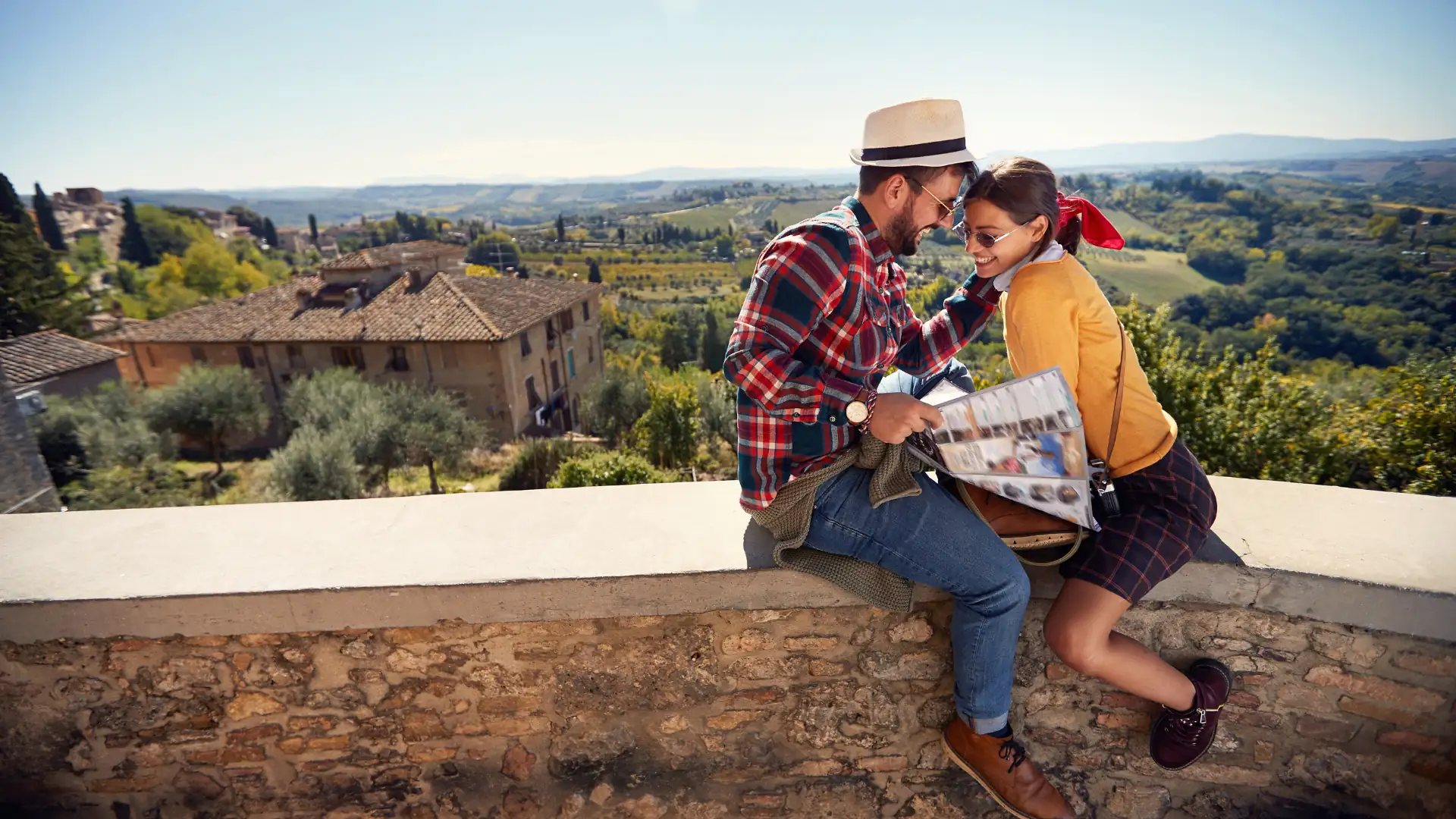 Paar sitzt auf einer Steinmauer mit Blick auf eine ländliche italienische Landschaft, der Mann trägt einen Strohhut und ein kariertes Hemd, die Frau ein rotes Haarband und eine gelbe Jacke.