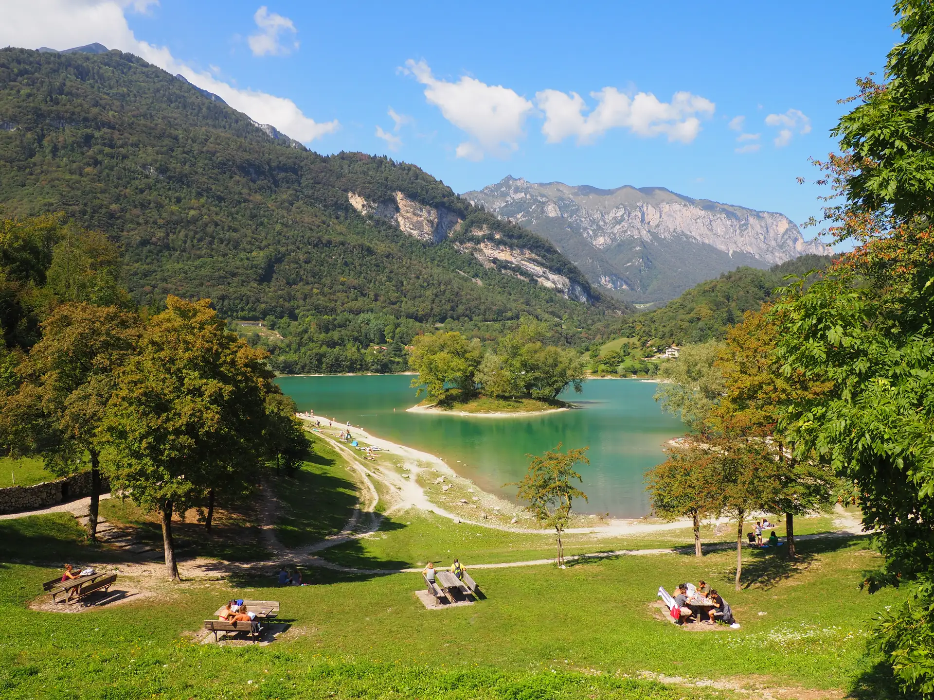 Grüne Wiese mit Picknicktischen und Menschen am Ufer des türkisfarbenen Tennosees umgeben von bewaldeten Bergen unter blauem Himmel
