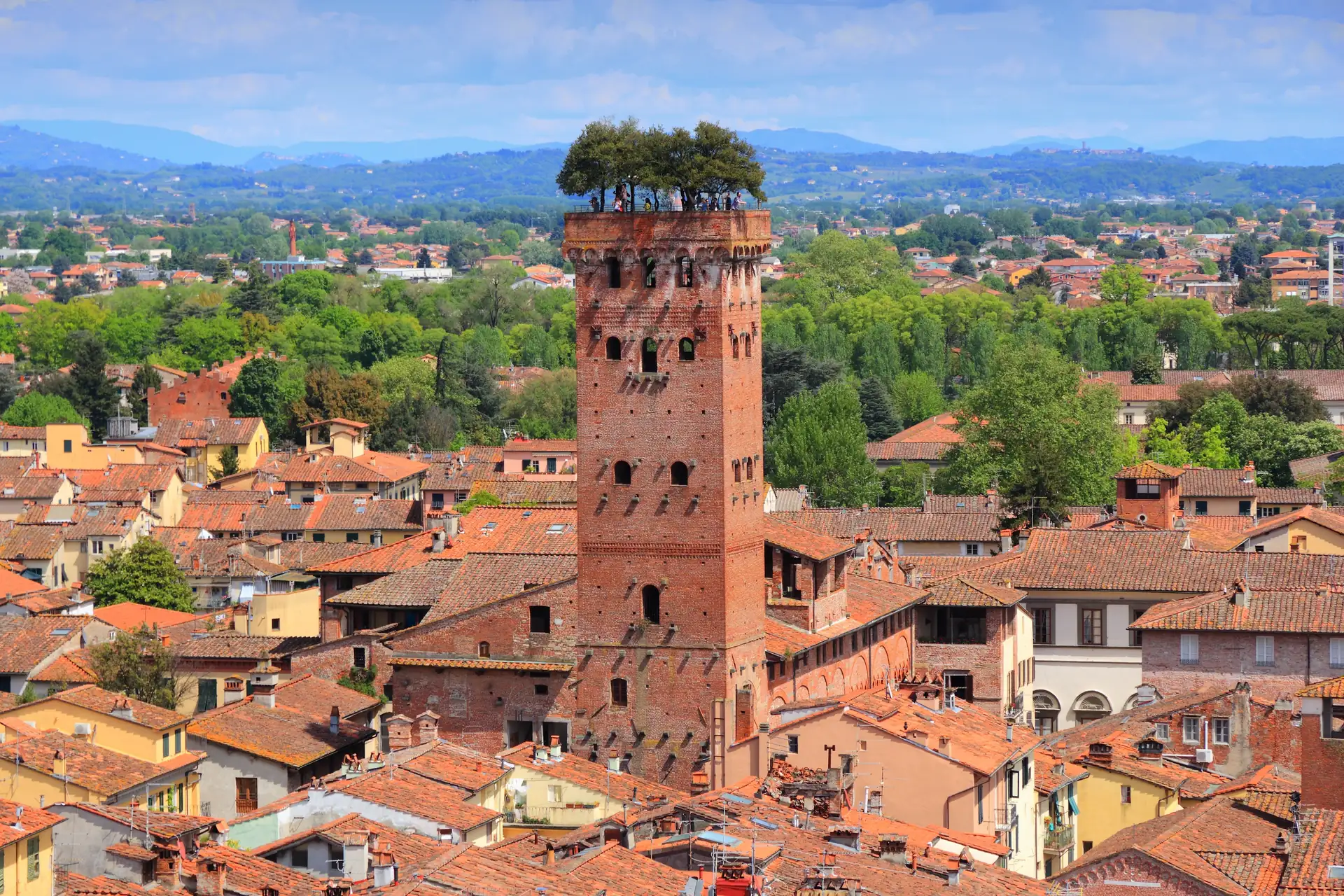 Roter Backsteinturm mit Bäumen auf der Spitze, umgeben von roten Ziegeldächern in der toskanischen Stadt Lucca mit grünen Hügeln im Hintergrund.
