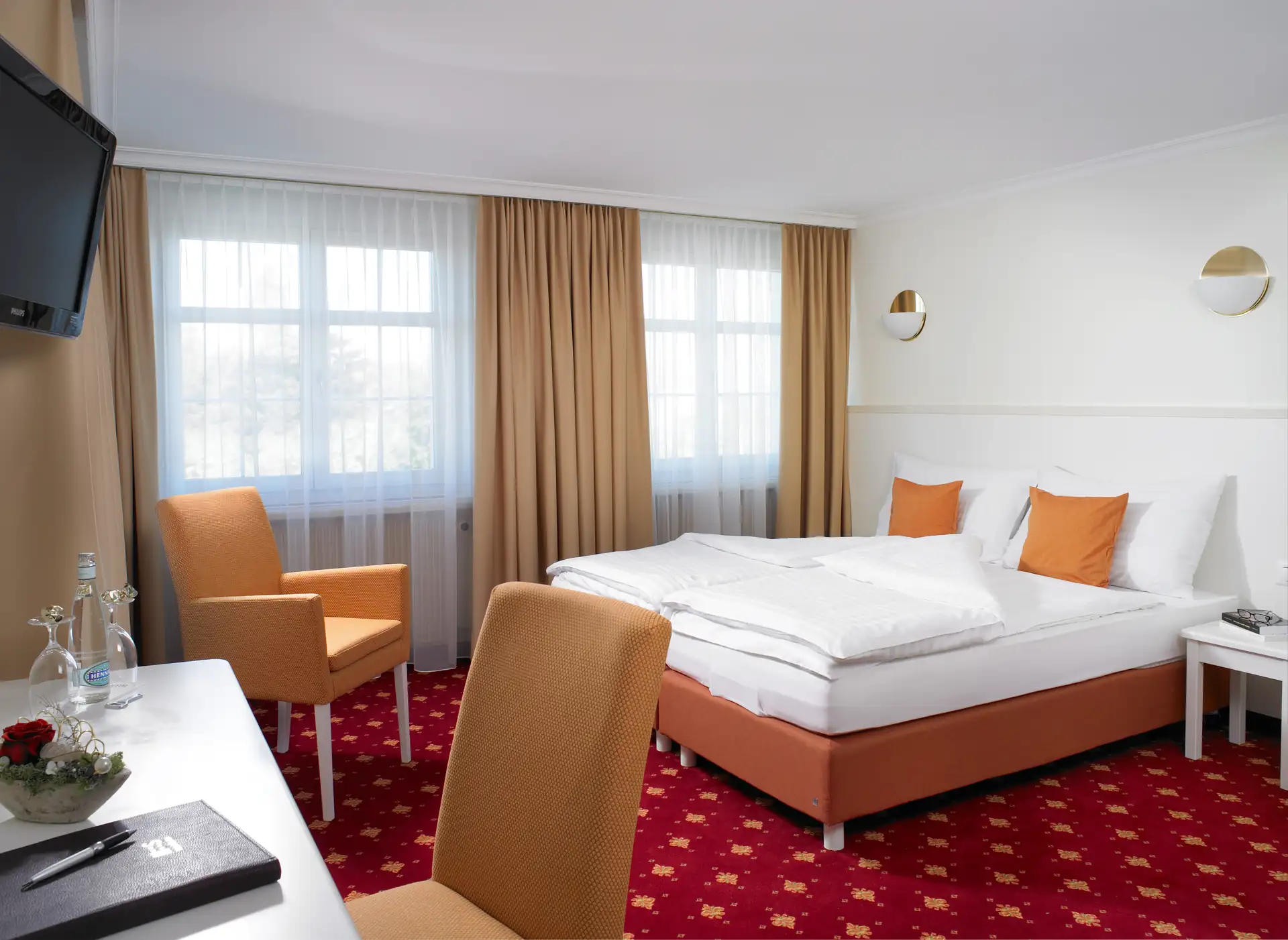Hotelzimmer mit Doppelbett, orangefarbenen Kissen und Vorhängen, rotem Teppich und Schreibtisch mit Stuhl im Hotel Swiss in Konstanz, Beispielhotel für die Radreise Bodensee