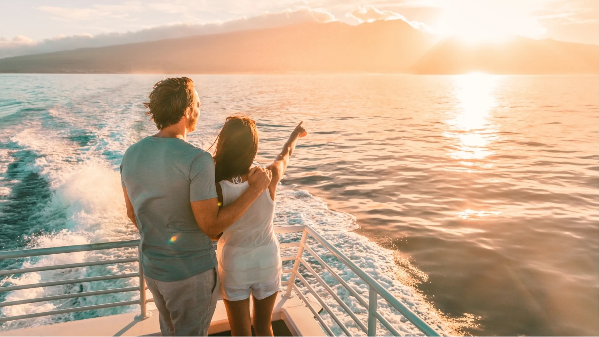 Ein Paar steht auf einem Boot, umarmt sich und blickt auf das Wasser und die untergehende Sonne. Die Frau trägt ein weißes Oberteil und Shorts, der Mann ein graues T-Shirt.