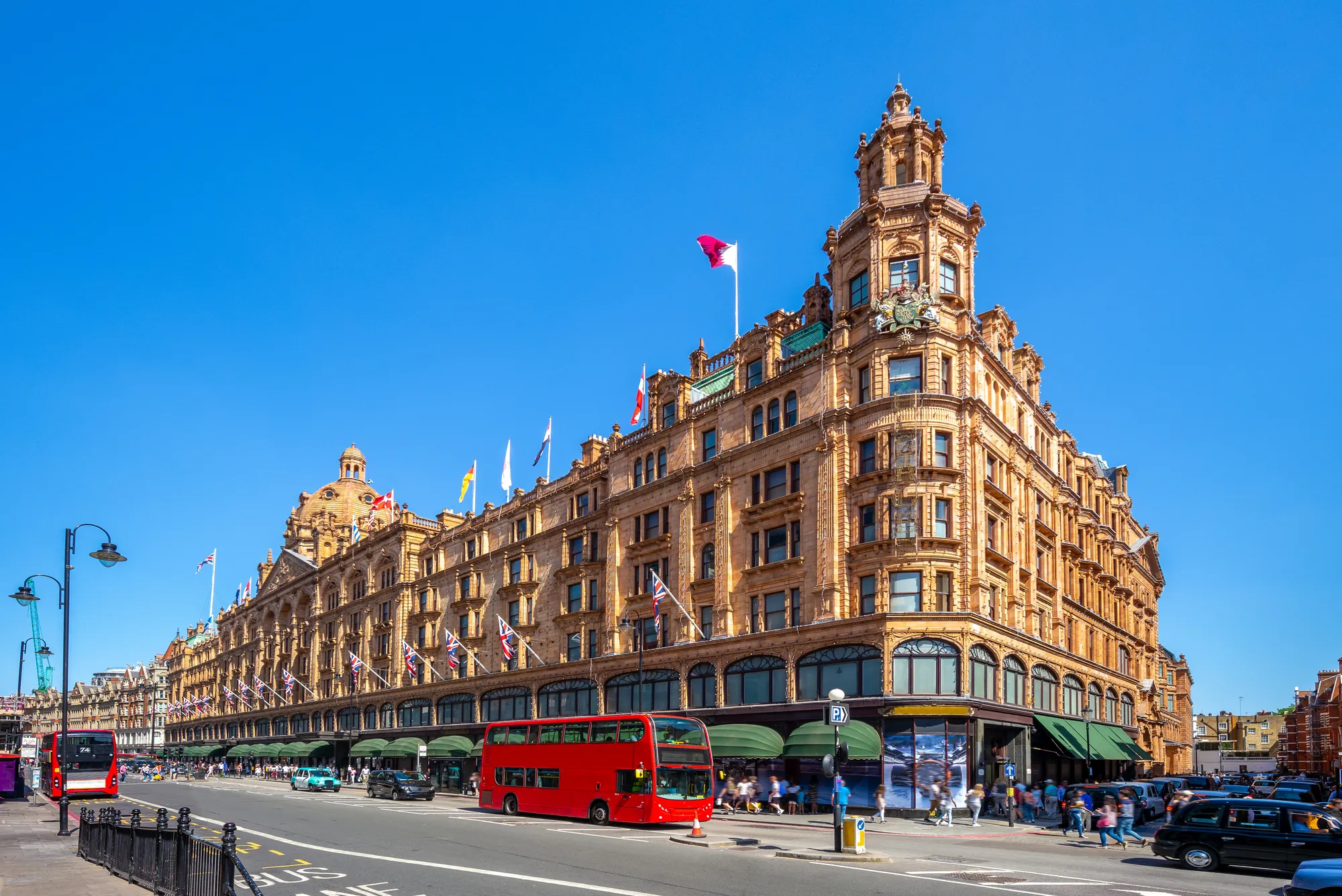 Das Bild zeigt Harrods, das weltberühmte Luxus-Kaufhaus in London. Das große, prächtige Gebäude mit seiner markanten viktorianischen Architektur erstreckt sich entlang der Brompton Road in Knightsbridge. Die Fassade ist mit vielen Flaggen geschmückt, die auf die internationale Bedeutung des Kaufhauses hinweisen. Vor dem Gebäude sind auch der ikonische rote Doppeldeckerbus und Londons schwarze Taxis zu sehen, was die typische Londoner Stadtkulisse widerspiegelt. Harrods ist bekannt für seine eleganten Schaufenster und seine breite Auswahl an luxuriösen Waren, die Touristen und Einheimische gleichermaßen anziehen.