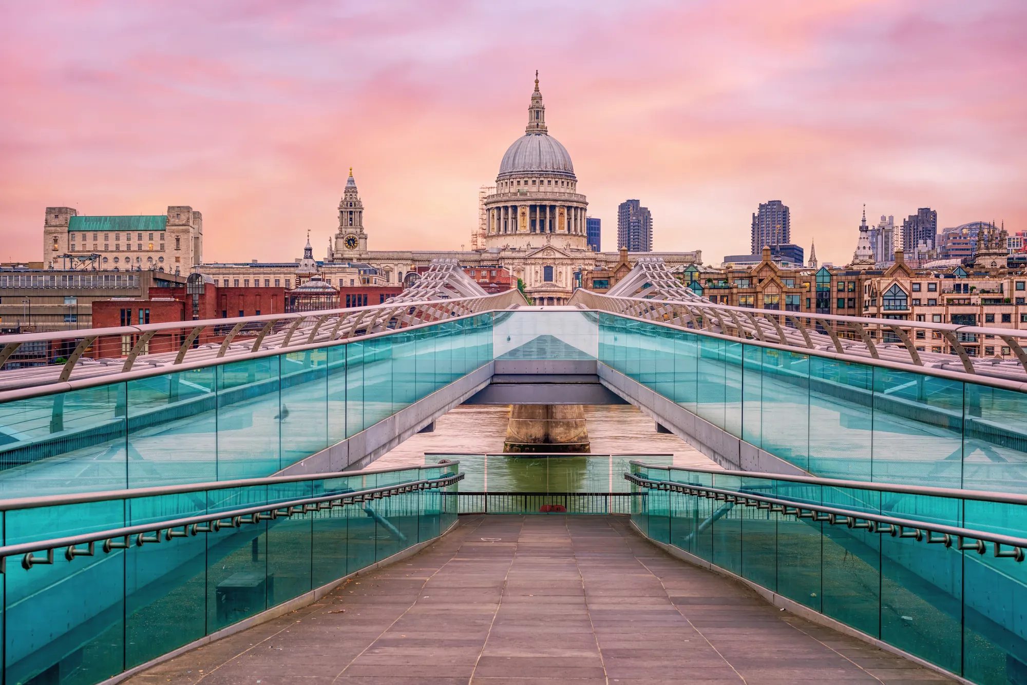 Das Bild zeigt einen Blick auf die ikonische St. Paul’s Cathedral in London, aufgenommen von der Millennium Bridge aus. Die architektonische Pracht der Kathedrale ragt über die Brücke hinaus, während die Brücke selbst mit ihren modernen Linien den Vordergrund dominiert. Der Himmel ist in sanften Pastelltönen gehalten, was der Szene eine ruhige und majestätische Atmosphäre verleiht. Die Mischung aus klassischer und moderner Architektur ist ein typisches Merkmal der Londoner Skyline.