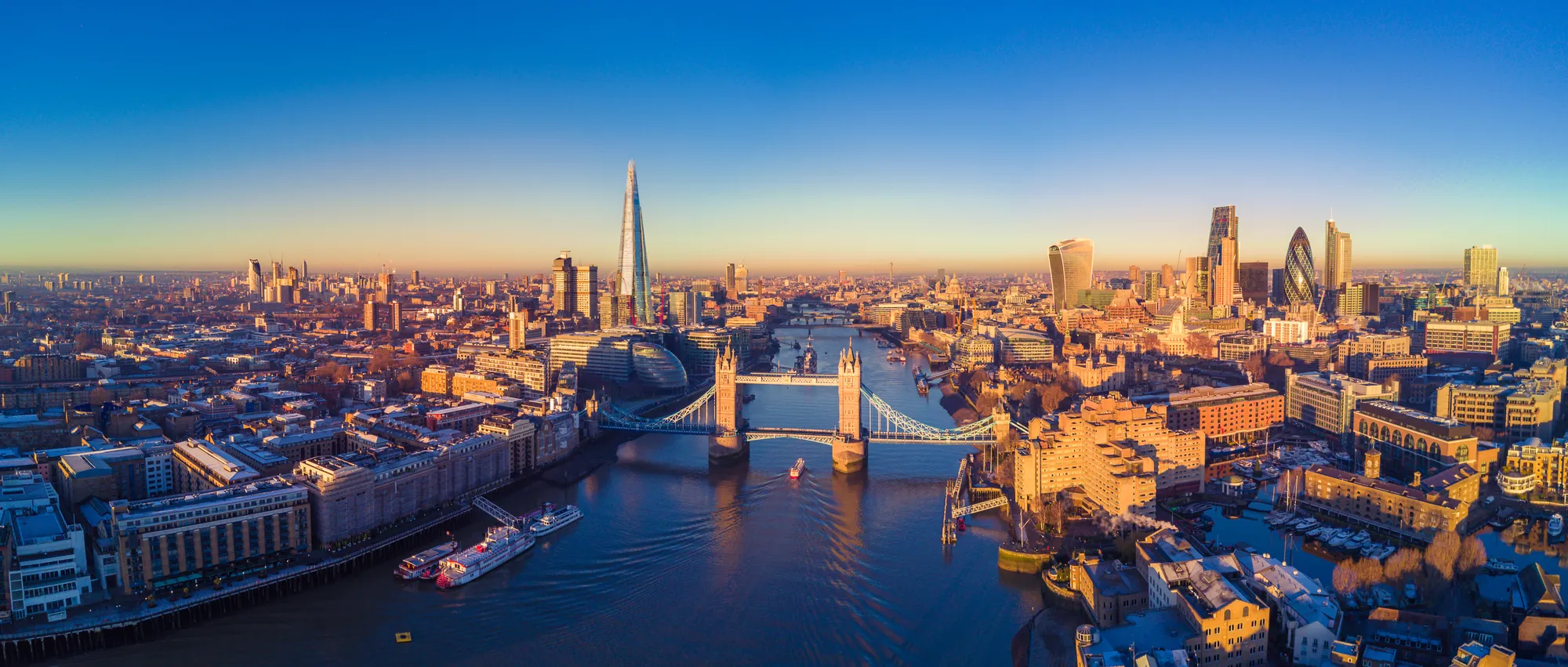Das Bild zeigt eine atemberaubende Luftaufnahme der Skyline von London, mit einem klaren Blick auf die berühmte Tower Bridge im Vordergrund. Im Hintergrund erhebt sich das moderne Londoner Stadtbild mit markanten Wolkenkratzern wie The Shard und The Gherkin. Der Fluss Thames schlängelt sich durch die Stadt und reflektiert die goldene Abendsonne, was der Szene eine warme und einladende Atmosphäre verleiht.