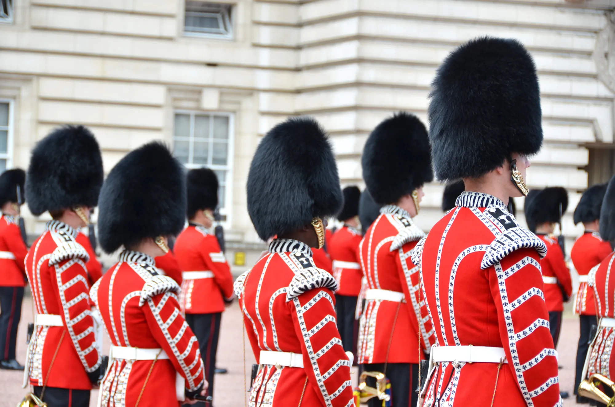 Das Bild zeigt die berühmte Wachablösung am Buckingham Palace in London. Die Soldaten der Queen's Guard tragen ihre ikonischen roten Uniformen und schwarzen Bärenfellmützen, während sie in geordneter Formation stehen. Diese Zeremonie, bei der die Wache am Palast wechselt, ist eine beliebte Touristenattraktion und symbolisiert die traditionsreiche Geschichte der britischen Monarchie.