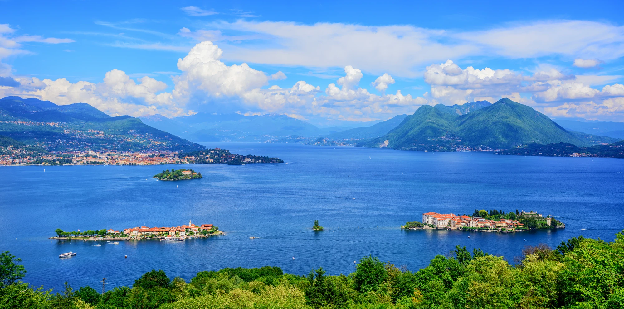Panorama des Lago Maggiore mit zwei Inseln in der Mitte, aufgenommen bei Tageslicht. Im Hintergrund erheben sich majestätische Berge und sanfte Landschaften, die zusammen ein harmonisches und malerisches Bild der Natur ergeben.