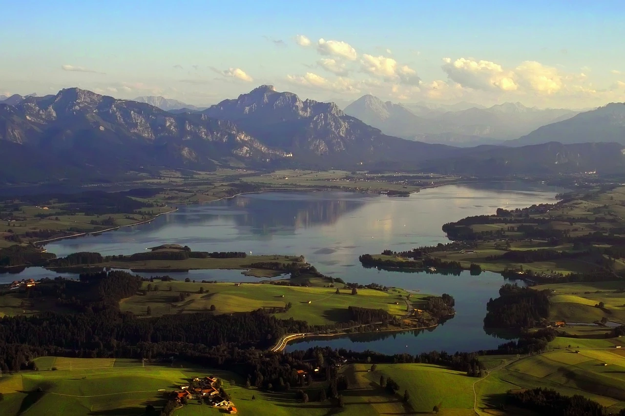 Das Bild zeigt den Forggensee in einer weitläufigen Ansicht, eingebettet in die sanfte Hügellandschaft des Allgäus und umgeben von den majestätischen Alpen. Die ruhige Wasseroberfläche spiegelt die Berge wider, während grüne Felder und kleine Ansiedlungen das Ufer säumen. Diese beeindruckende Panoramaaufnahme vermittelt eine friedliche Atmosphäre und zeigt die Schönheit der bayerischen Voralpenlandschaft in ihrer ganzen Pracht.