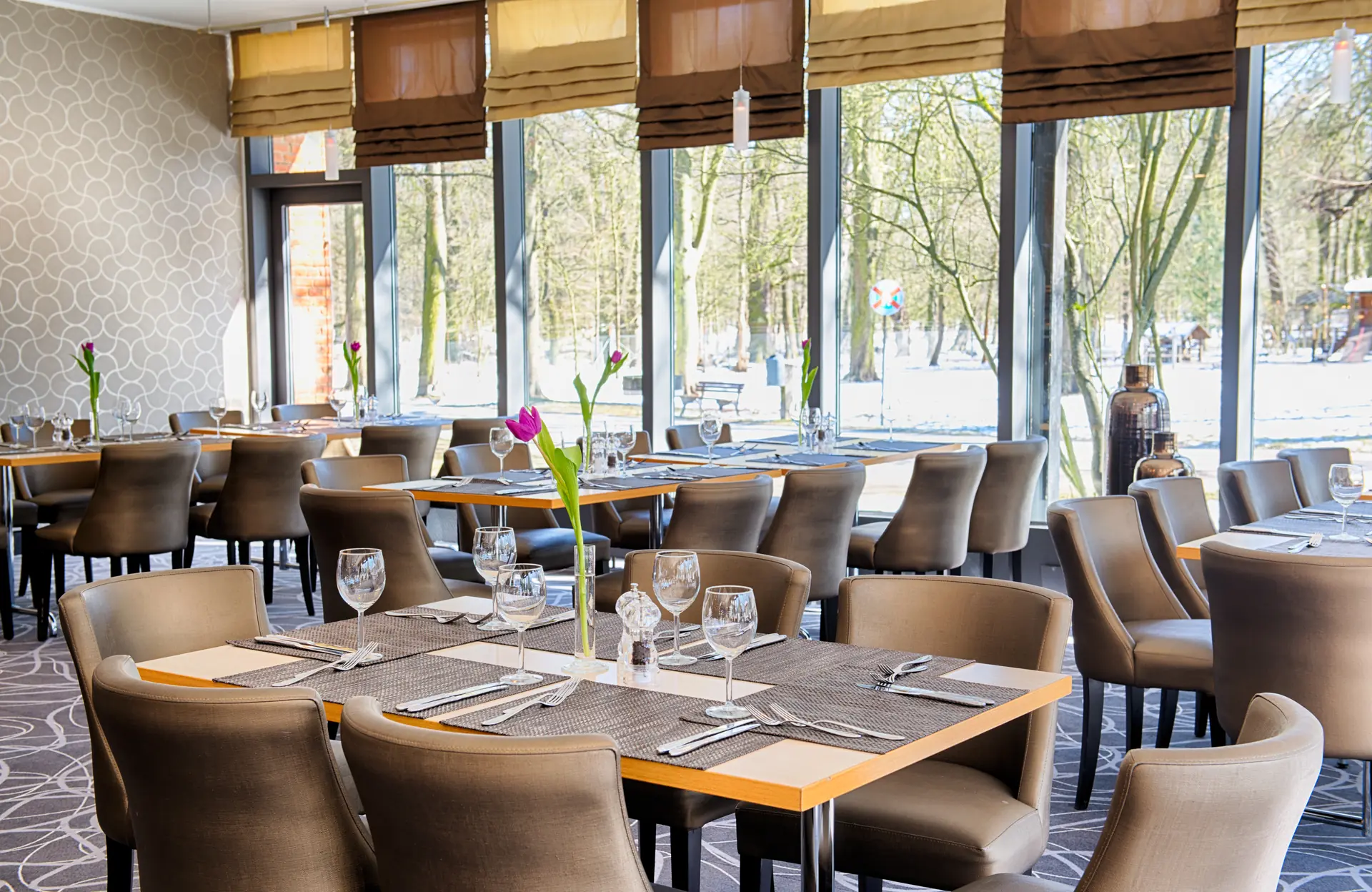 Eingedeckte Tische im Restaurant des Leonardo Hotels Hannover mit großer Fensterfront, die auf den benachbarten Tiergarten blicken lässt