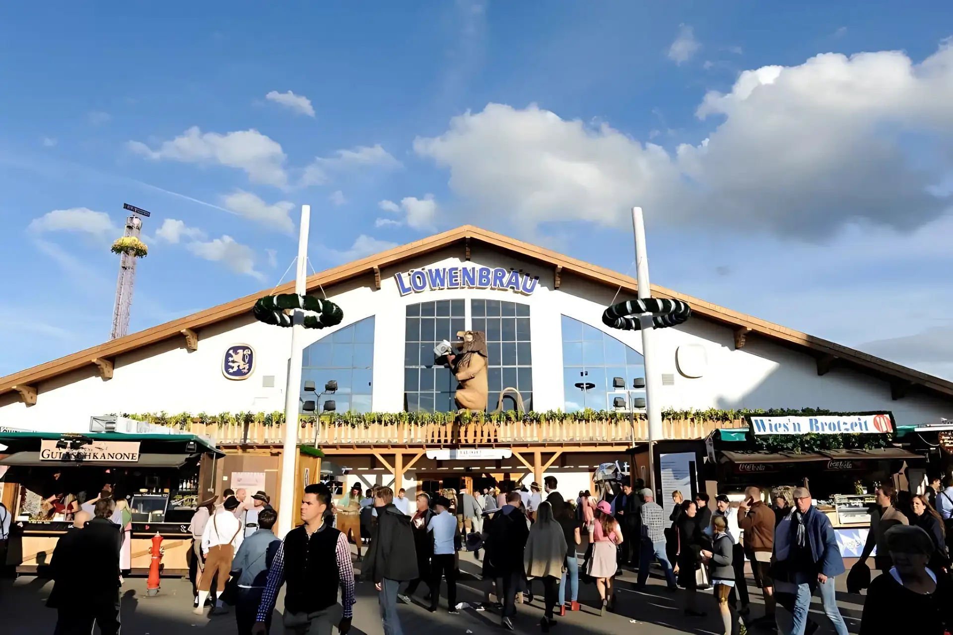 Löwenbräu Festzelt auf dem Münchner Oktoberfest: Besucher strömen in das Festzelt bei Sonnenschein mit Löwenfigur über dem Eingang im Fokus.