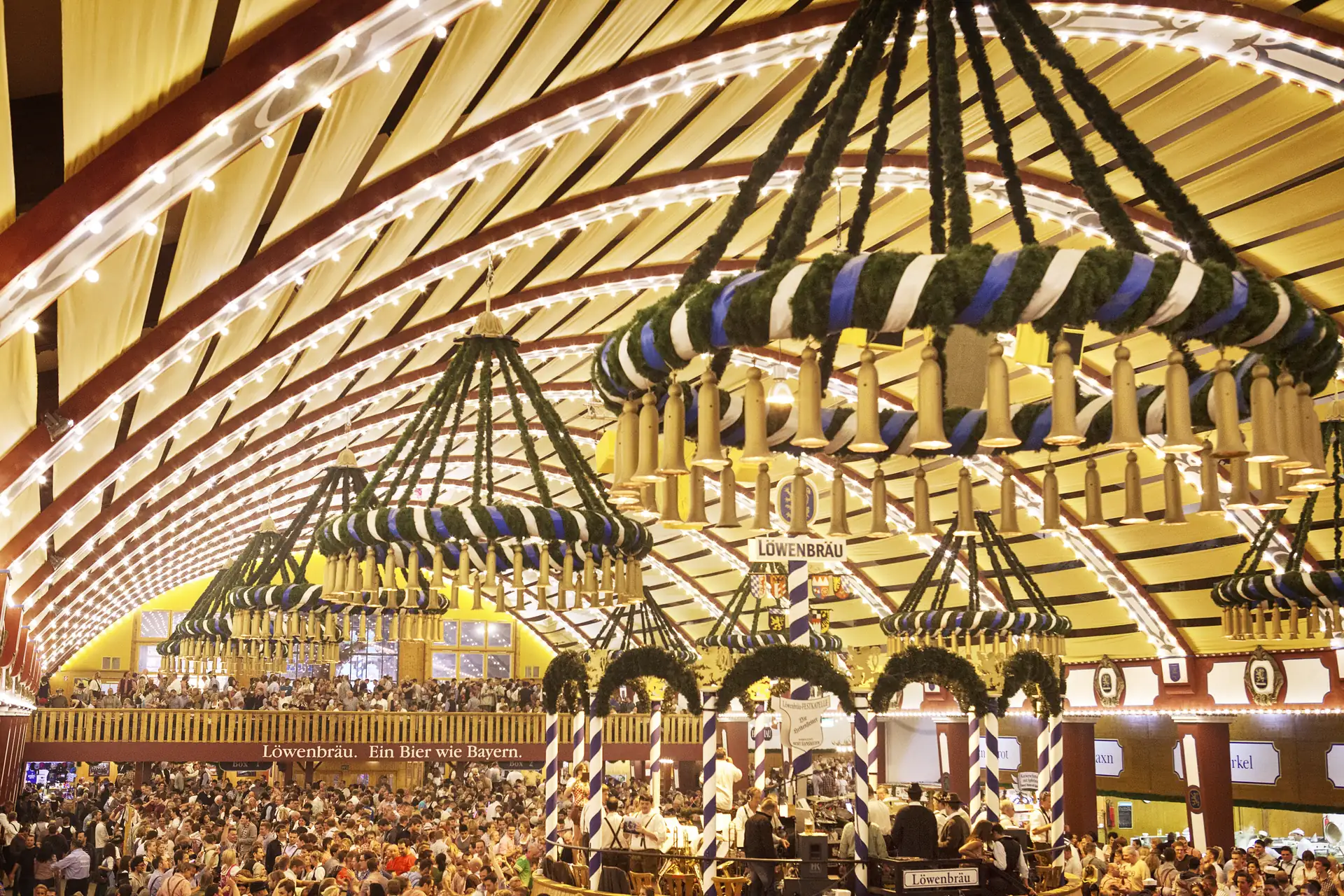 Löwenbräu Festzelt von innen: Große Kränze hängen von der Festzeltdecke, Blick auf die Musikbühne, die zahlreichen Festzeltbesucher sowie die Empore/Balkon im Hintergrund.