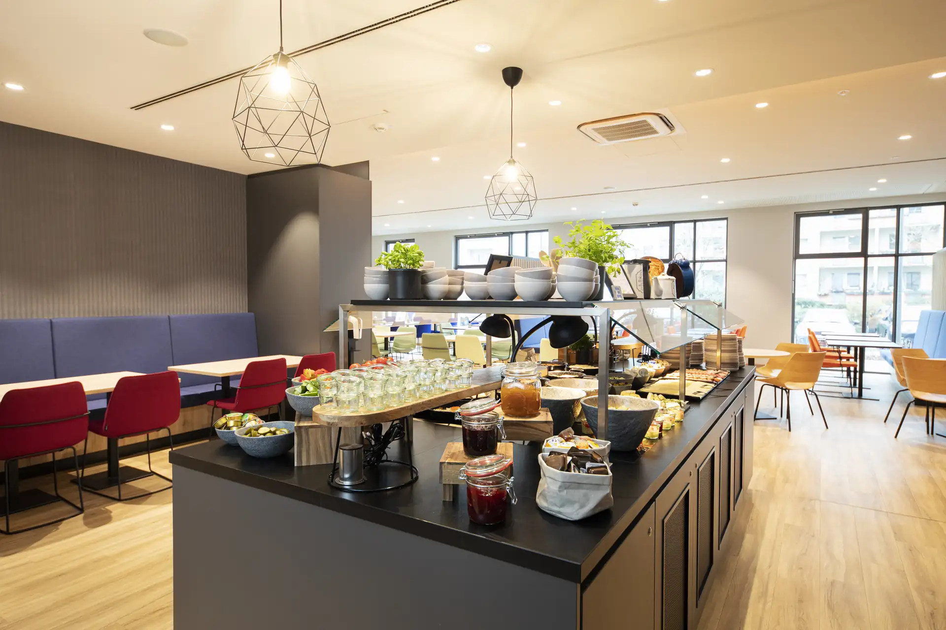 Loginn Hotel Berlin Airport: Buffetbereich mit verschiedenen Speisen und Getränken, darunter Obst, Joghurt und Marmelade. Sitzgelegenheiten im Hintergrund.