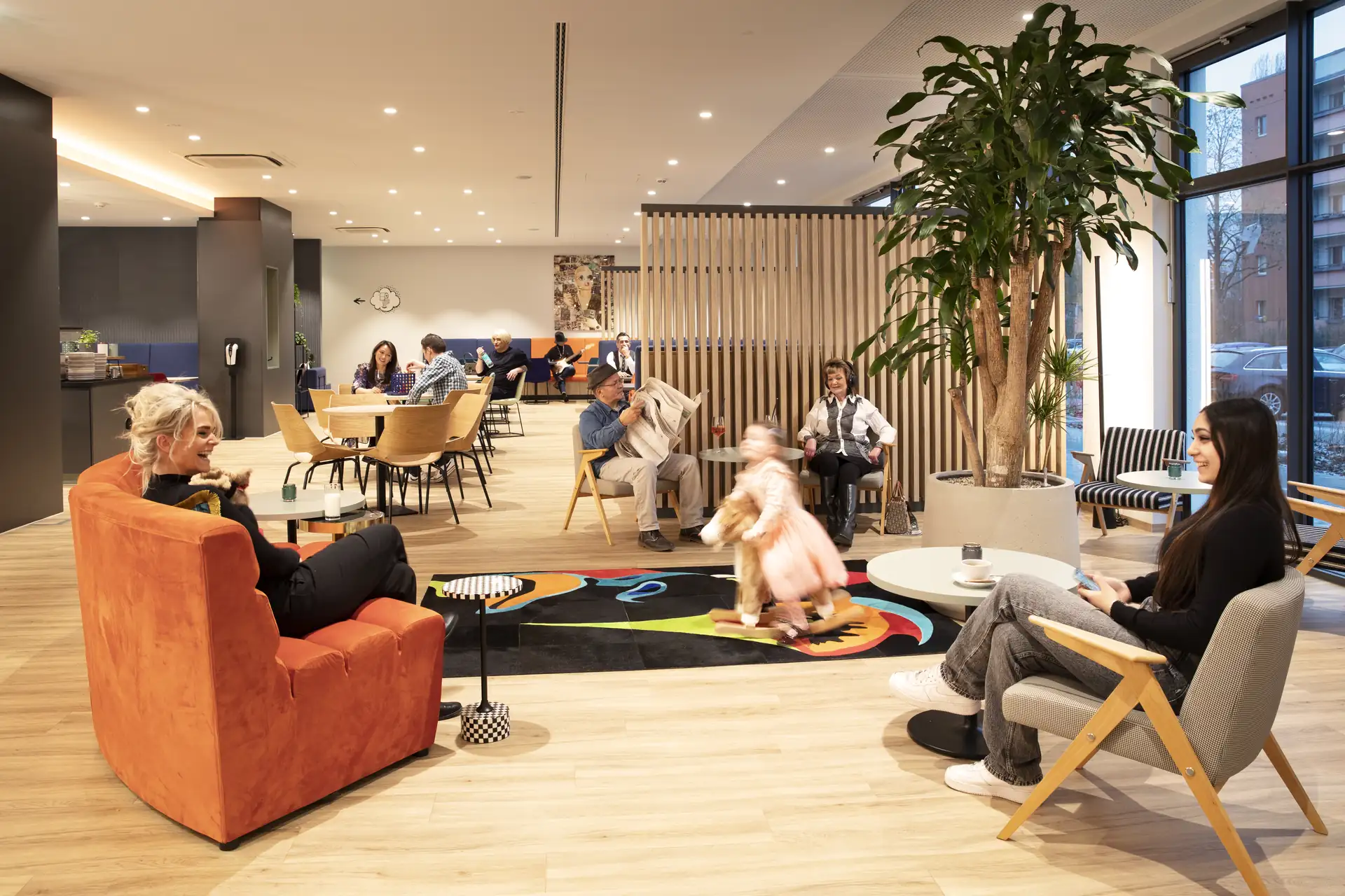 Loginn Hotel Berlin Airport: Lobby mit mehreren Sitzbereichen. Eine orangefarbene Couch, ein Teppich mit buntem Muster, Pflanzen und Gäste, die sich unterhalten.