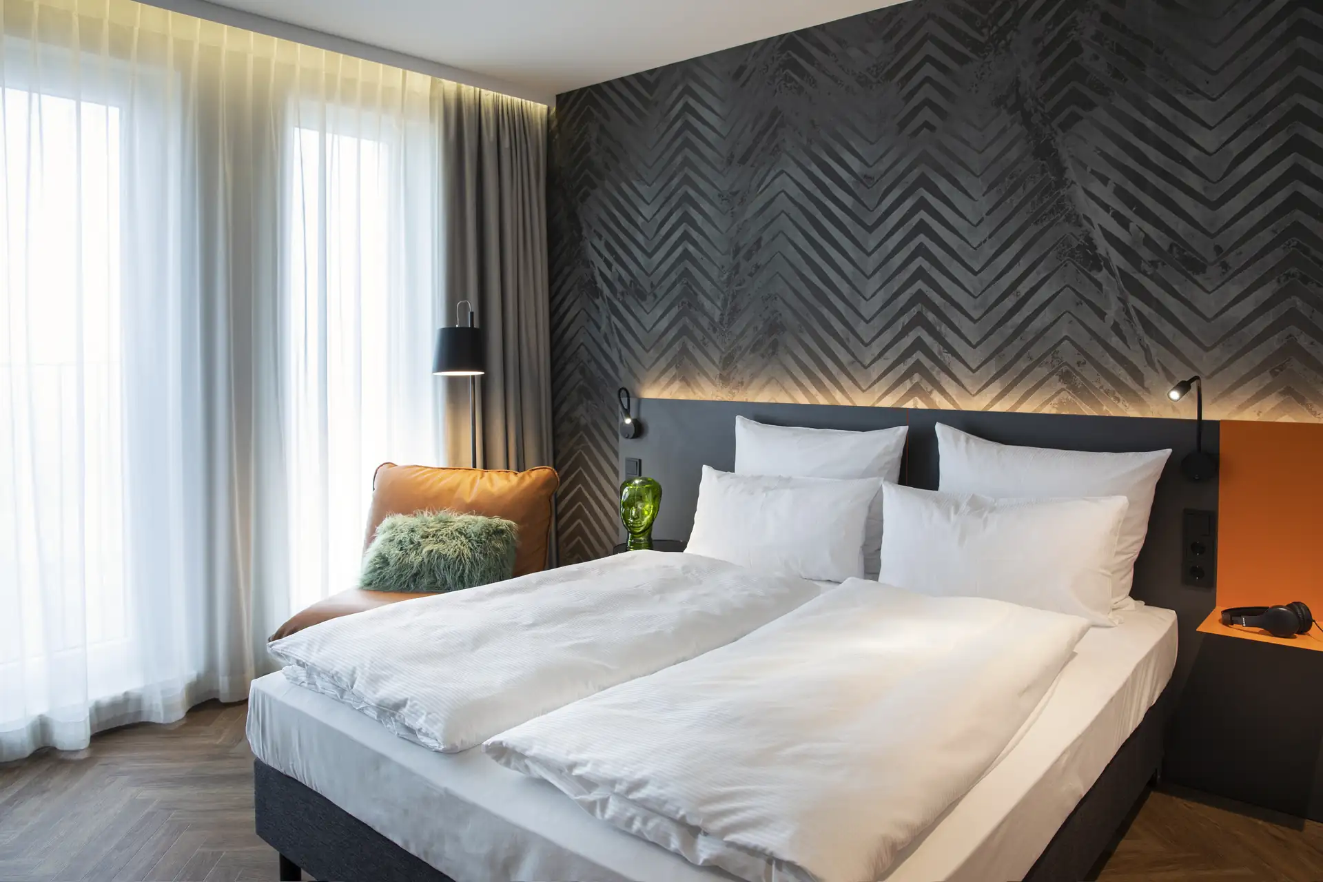 Loginn Hotel Berlin Airport: Doppelbett mit weißen Bettdecken in einem modernen Hotelzimmer, graue Wand mit chevron-Muster, große Fenster mit Vorhängen, orangefarbener Sessel.