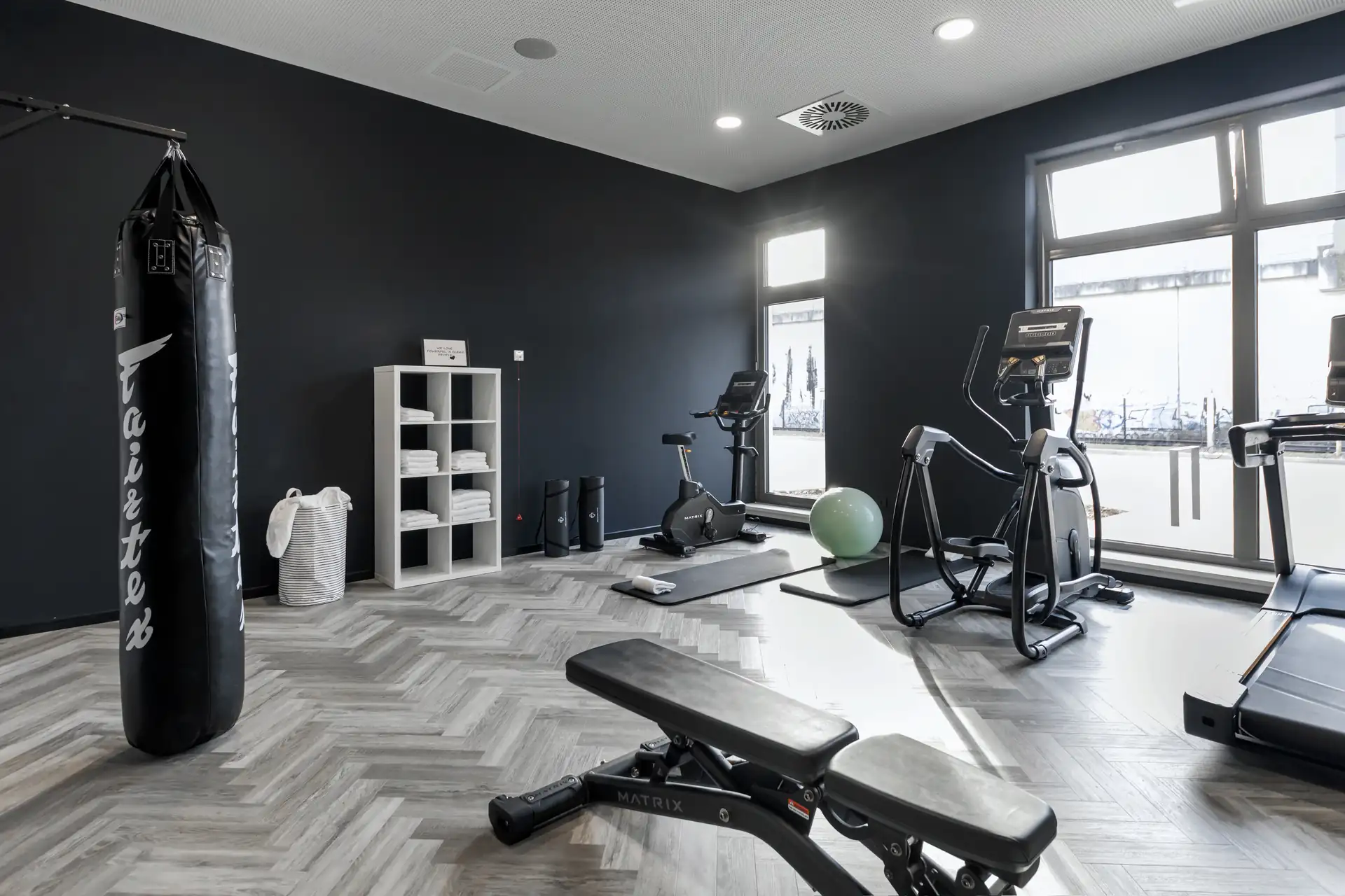 Loginn Hotel Köln Airport: Fitnessraum mit einem Boxsack, einem Laufband, einem Crosstrainer, einer Hantelbank und einem Gymnastikball. Helle Wände und große Fenster.