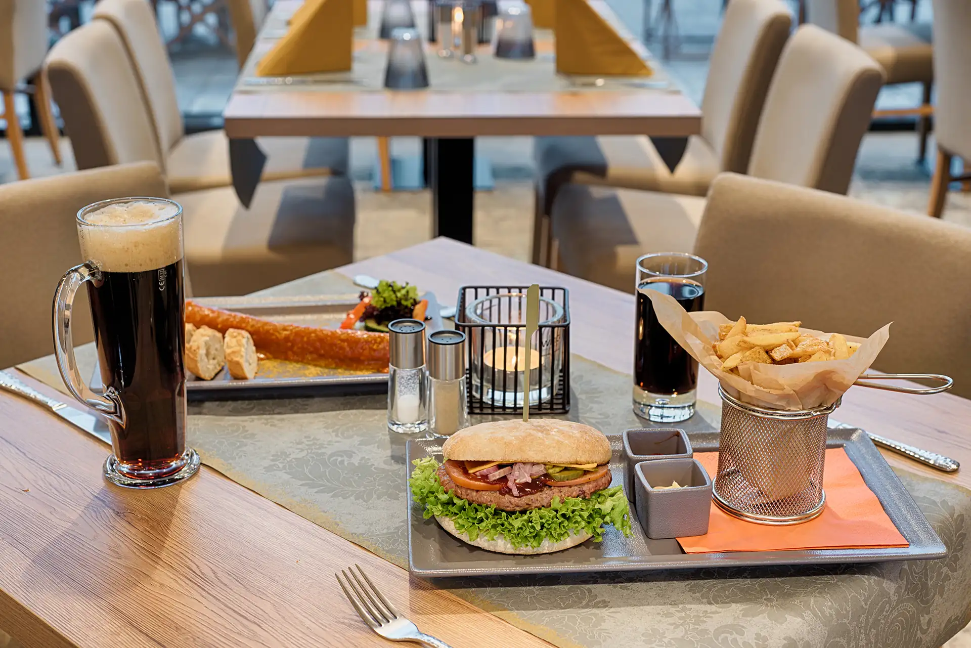 Tisch im Restaurant des Loginn Hotels Leipzig mit einem Burger auf Salat, Pommes in einem kleinen Drahtkorb, zwei Gläsern dunklem Bier und einem Teller mit Brot und Butter.
