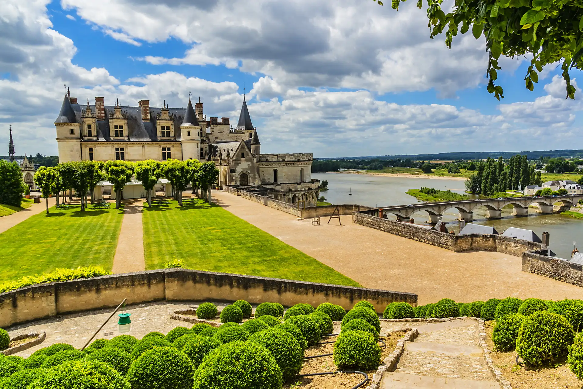 Schloss Amboise mit Türmen und Zinnen an der Loire, davor gepflegter Garten mit runden Buchsbaumhecken und Baumreihe, im Hintergrund Fluss und Brücke unter bewölktem Himmel.