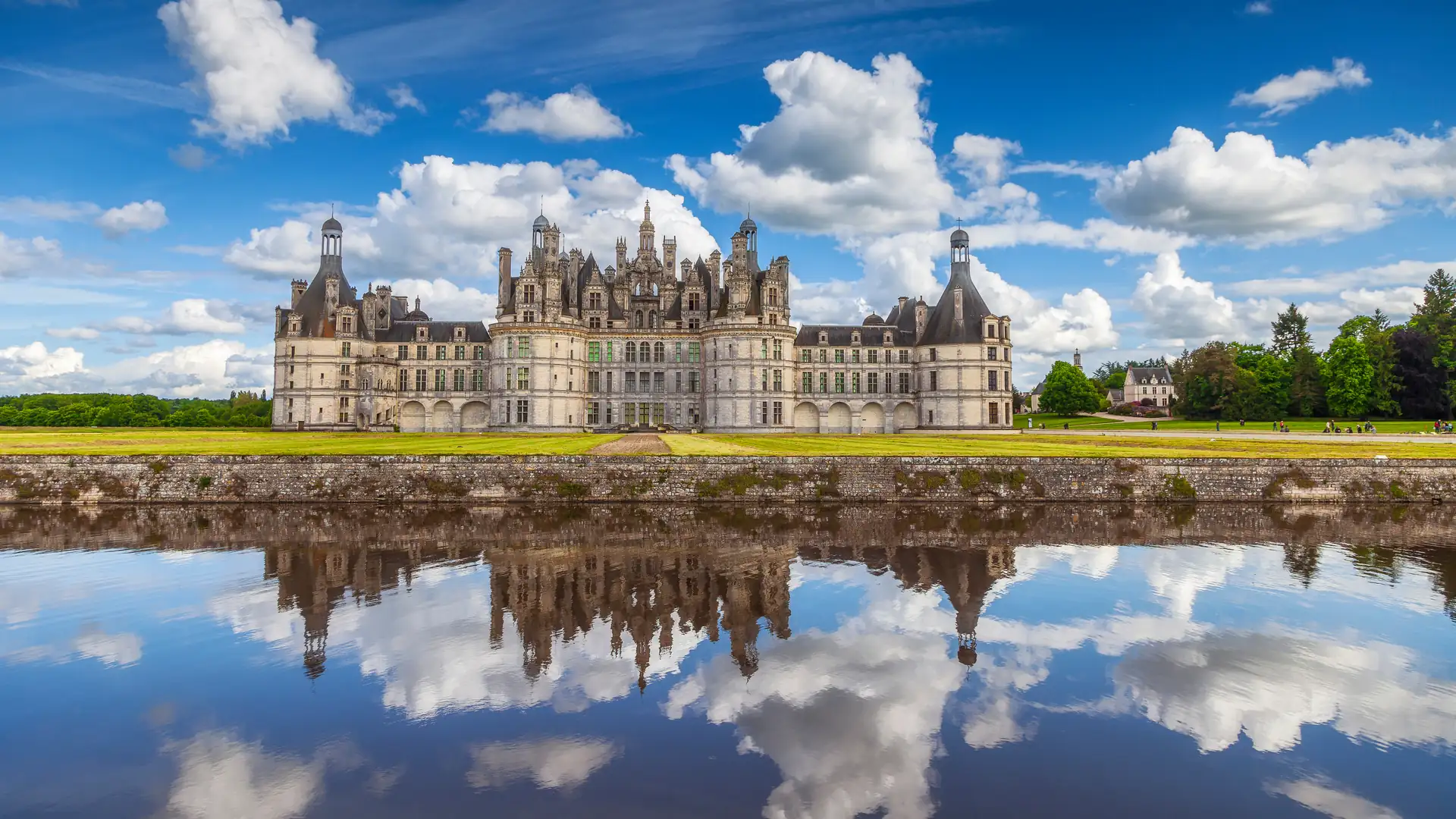 Schloss Chambord mit zahlreichen Türmen und Zinnen, spiegelnd im Wasser unter blauem Himmel mit weißen Wolken.