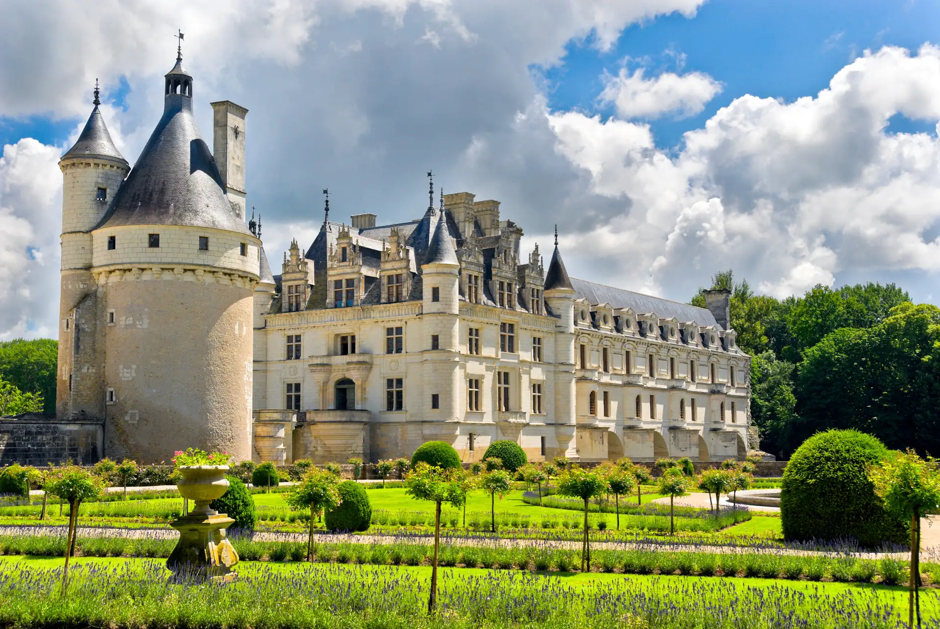 Schloss Chenonceau mit runden Türmen und symmetrischem Garten mit Hecken und Bäumen unter bewölktem Himmel.