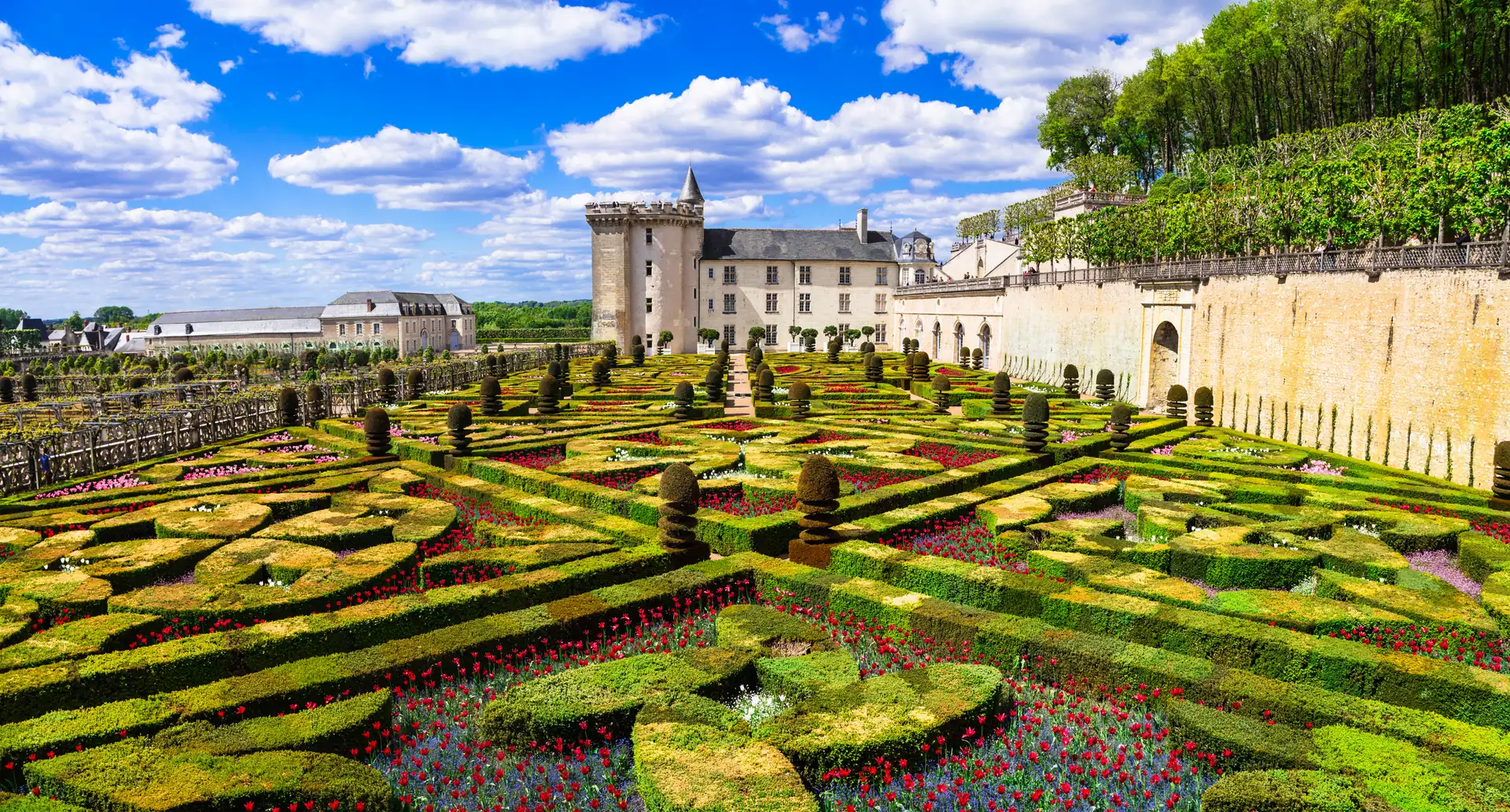 Symmetrisch angelegte Gartenanlage mit kunstvoll geschnittenen Hecken und roten Blumen vor dem historischen Schloss Villandry unter blauem Himmel mit Wolken.