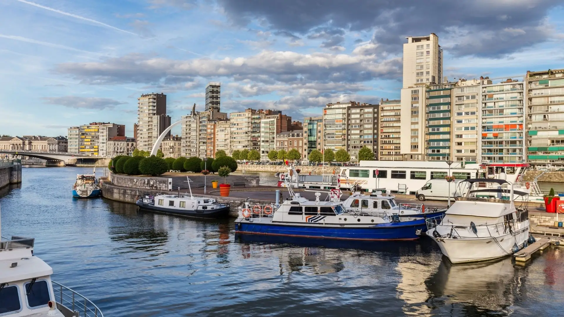 Blick auf den Hafen von Lüttich mit mehreren Booten am Ufer und Hochhäusern im Hintergrund unter bewölktem Himmel.