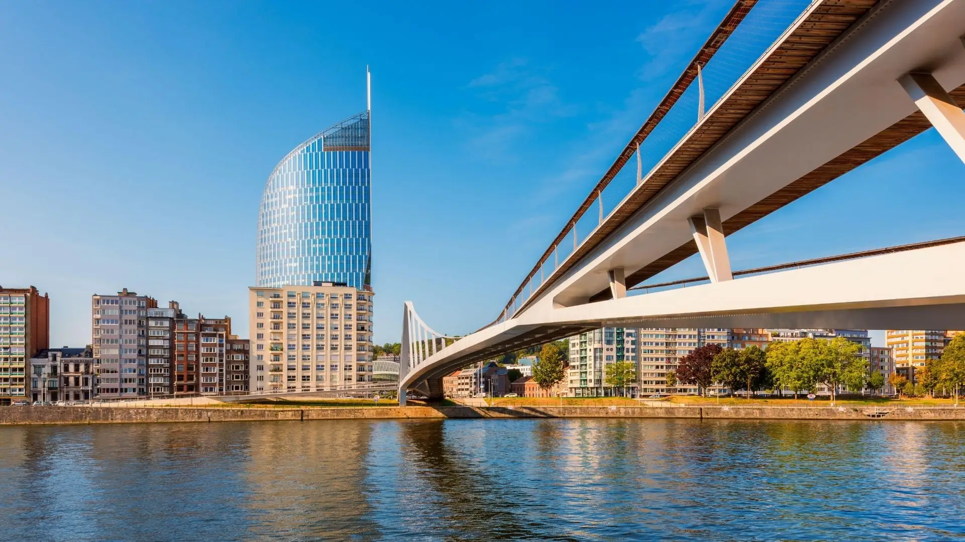 Blick auf die moderne Hochhausfassade und eine geschwungene Brücke über den Fluss in Lüttich bei klarem Himmel.