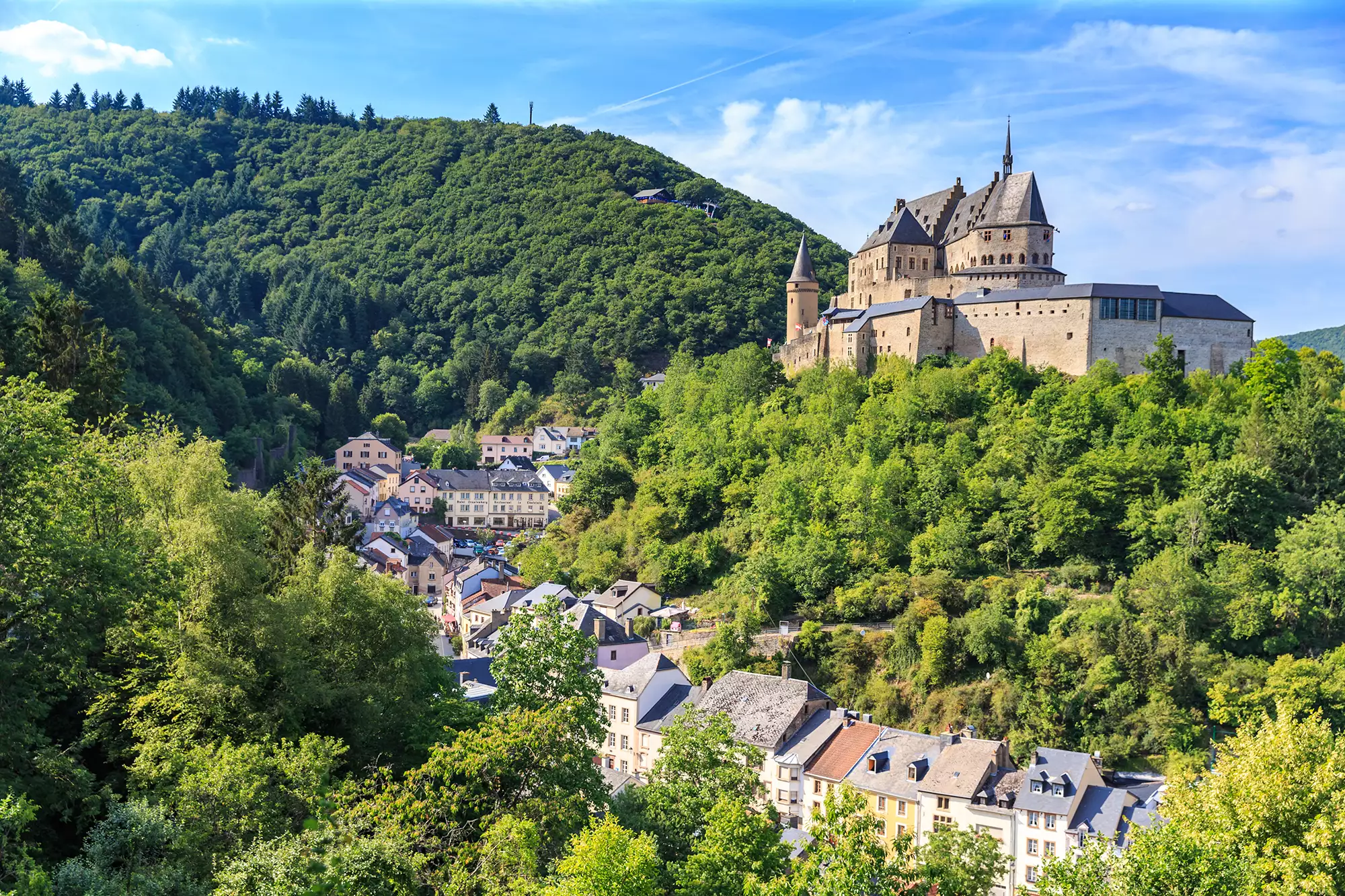 Blick auf die von Bäumen umgebene Burg Vianden bei klarem Wetter. Die majestätische Architektur der Burg harmoniert perfekt mit der umgebenden Natur und bietet ein beeindruckendes Bild.