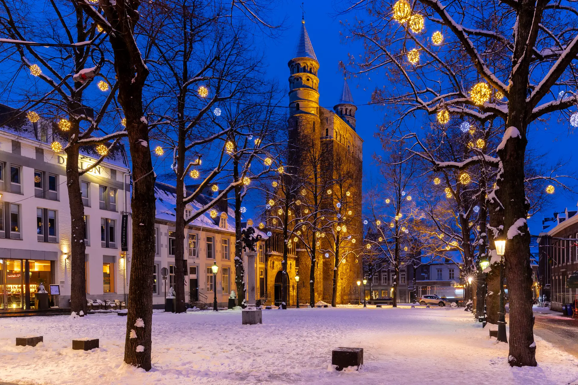 Weihnachtlich beleuchteter Platz in Maastricht mit schneebedecktem Boden, Bäumen und einer historischen Kirche im Hintergrund bei Abenddämmerung.
