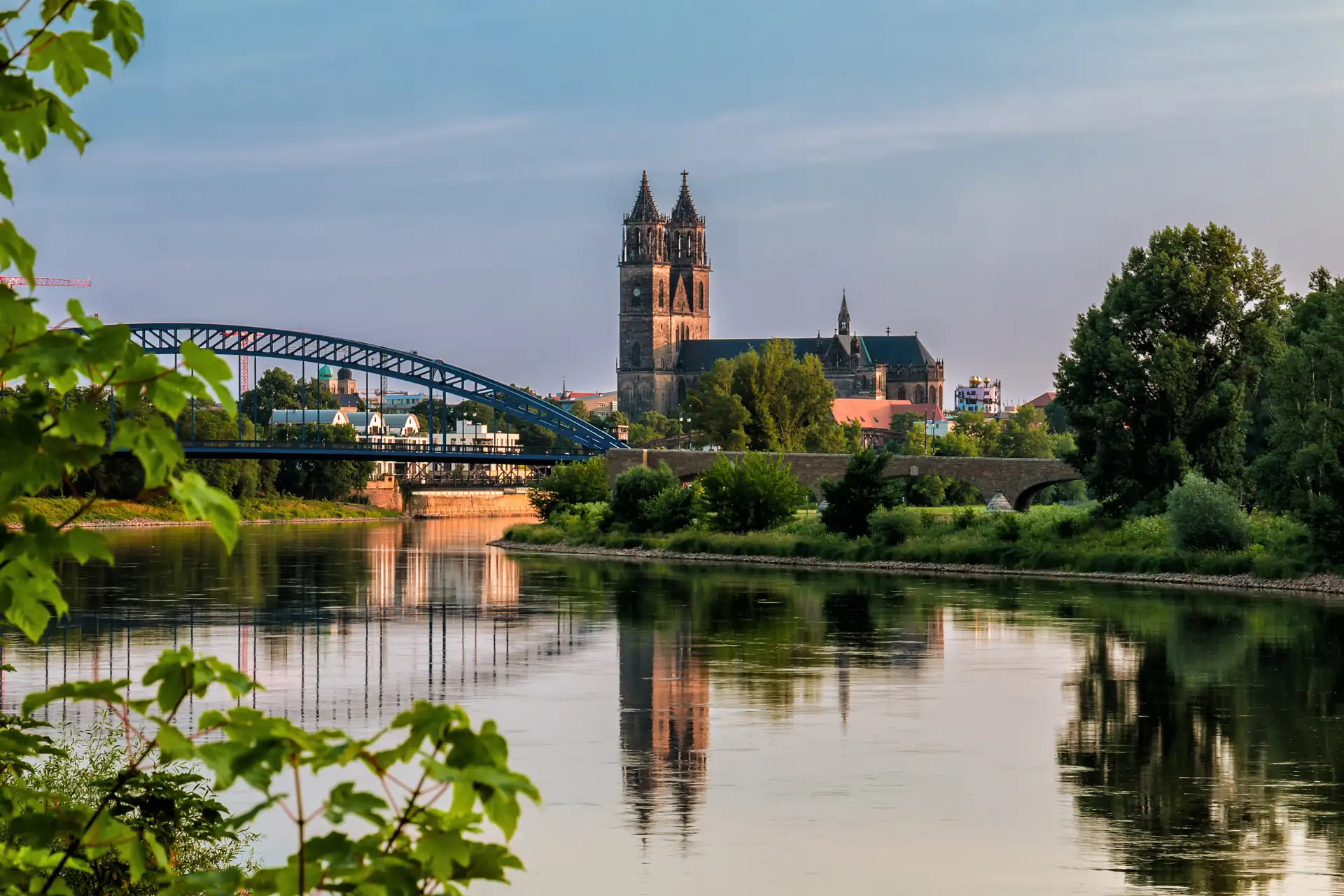 Blick auf den Magdeburger Dom mit zwei Türmen und eine Brücke über einen Fluss, umgeben von Bäumen und grüner Vegetation