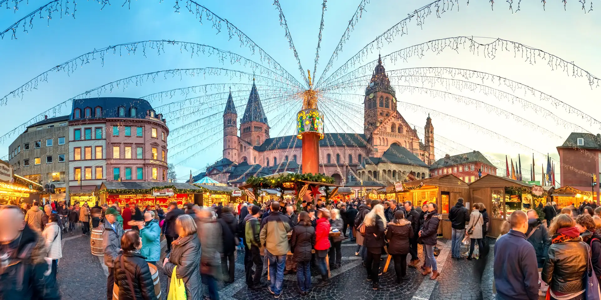 Mainzer Weihnachtsmarkt mit vielen Besuchern, beleuchteten Ständen und einer großen Pyramide vor dem Mainzer Dom bei Abenddämmerung.