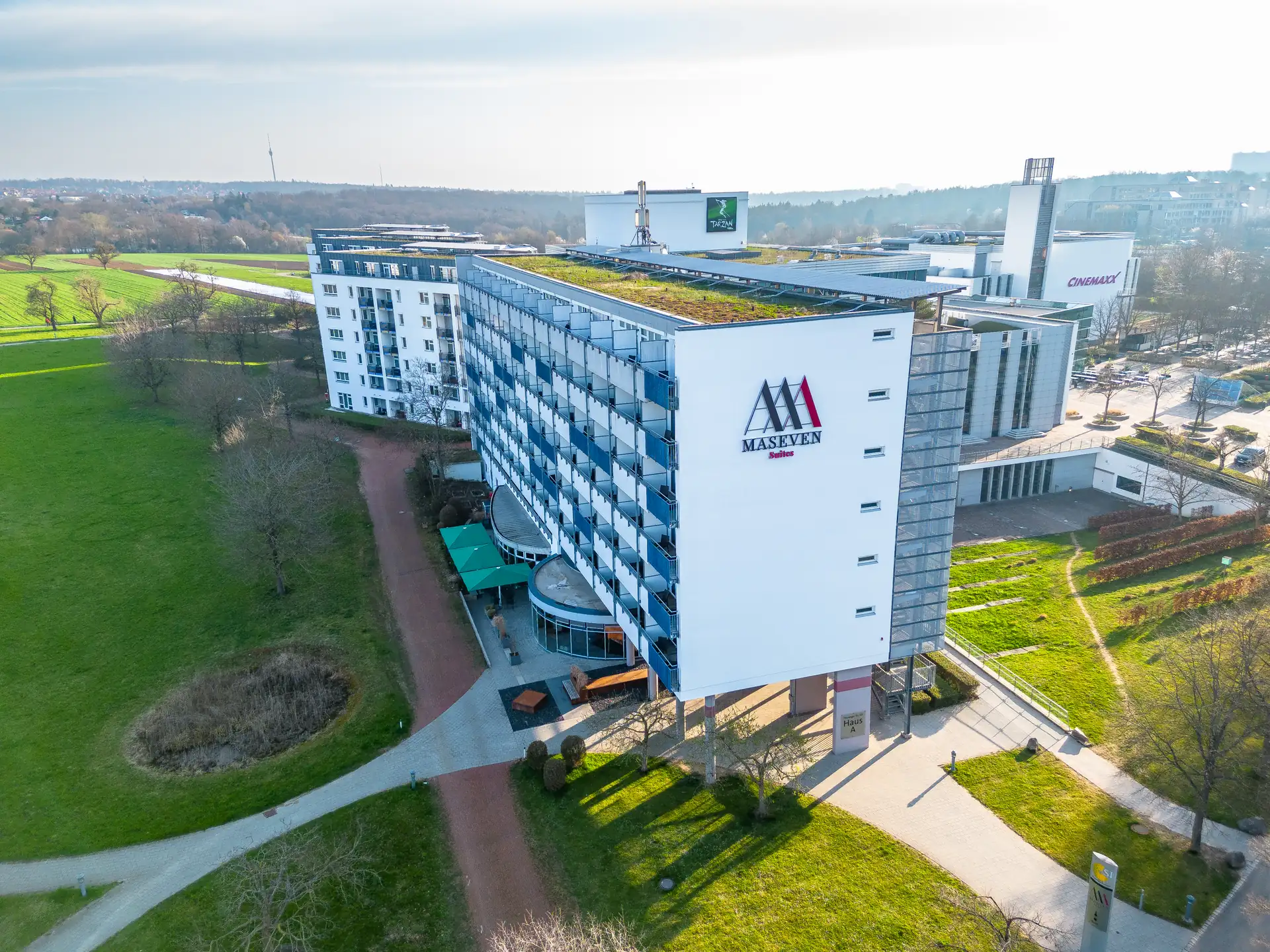 MASEVEN Suites in Stuttgart: Modernes Hotelgebäude mit grünem Dach und Logo 'MASEVEN Suites' an der Fassade, umgeben von Grünflächen und Wegen