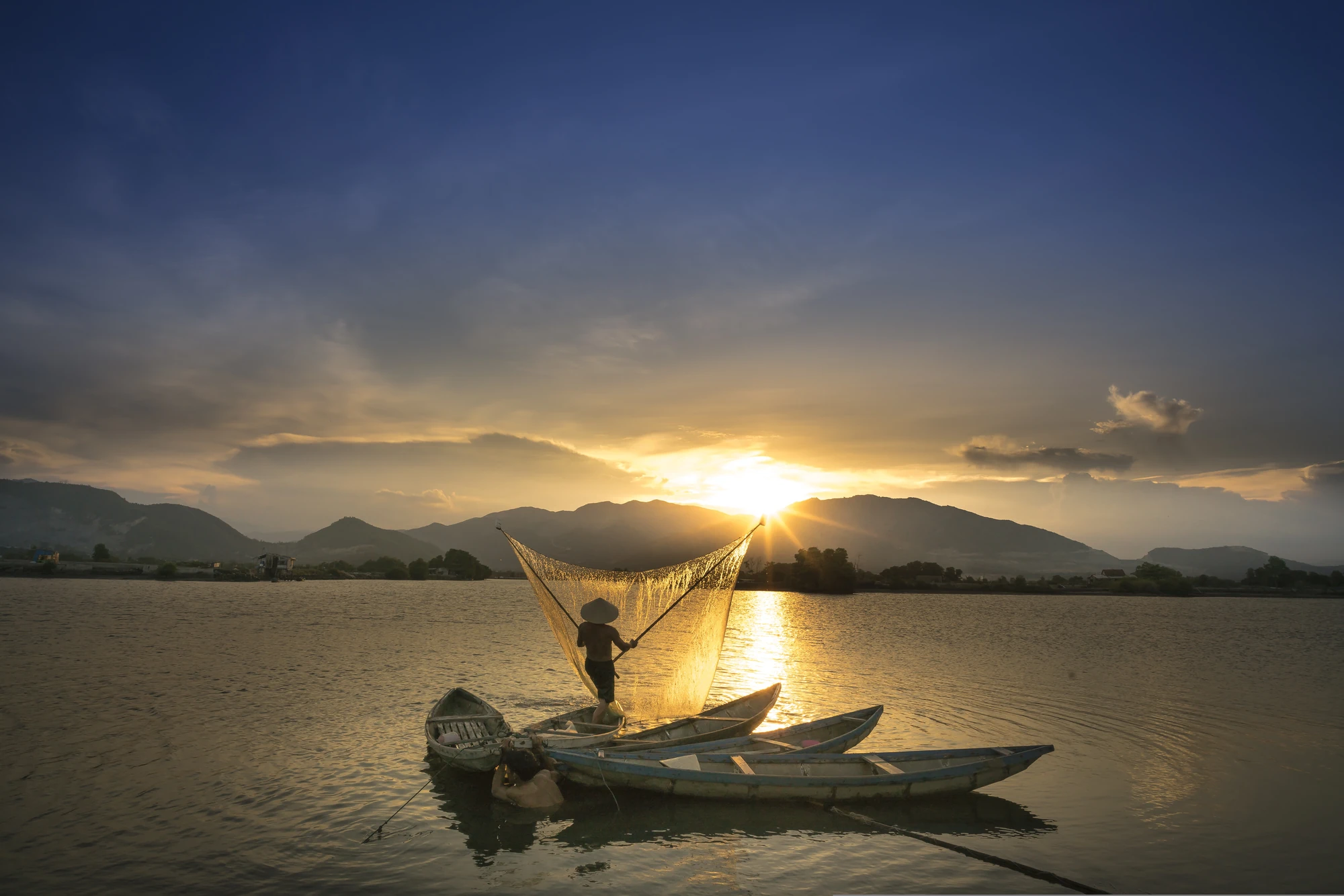 Ein Fischer mit einem Netz steht auf einem Boot im Mekong bei Sonnenuntergang. Im Hintergrund sind Berge und ein farbenfroher Himmel sichtbar.